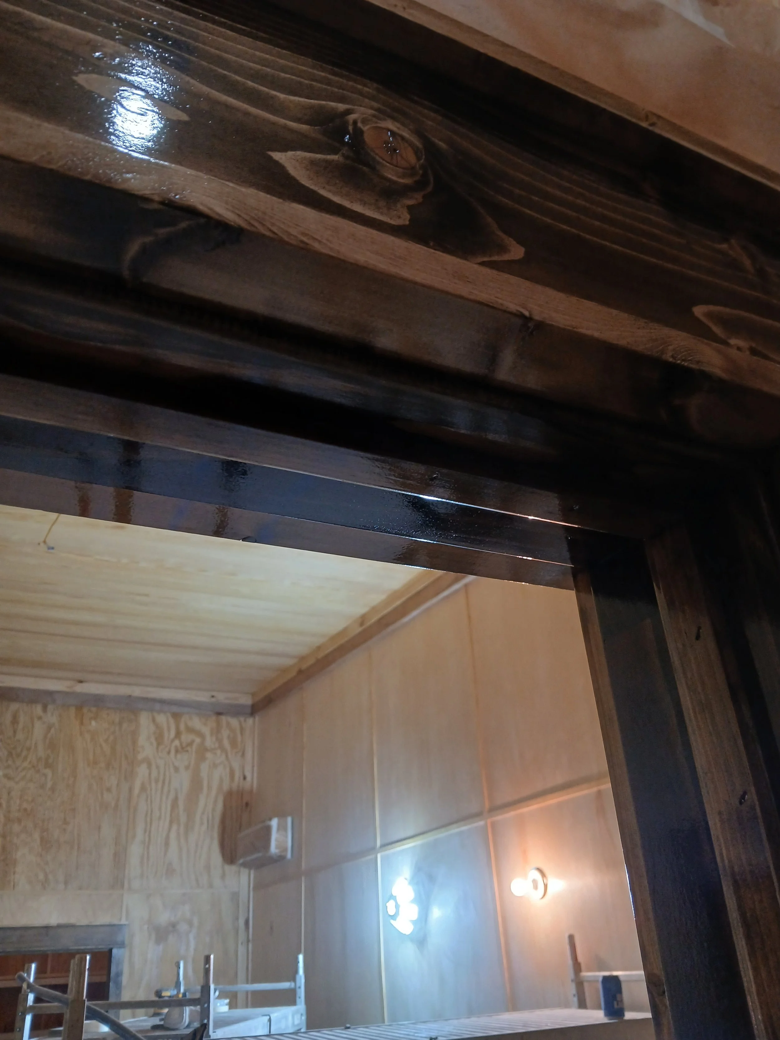 Close-up view of a wooden structure with glossy, dark brown finish, underneath a ceiling with light-colored wood paneling, in a room with tiled walls, a wall-mounted light fixture, and a metal ladder.