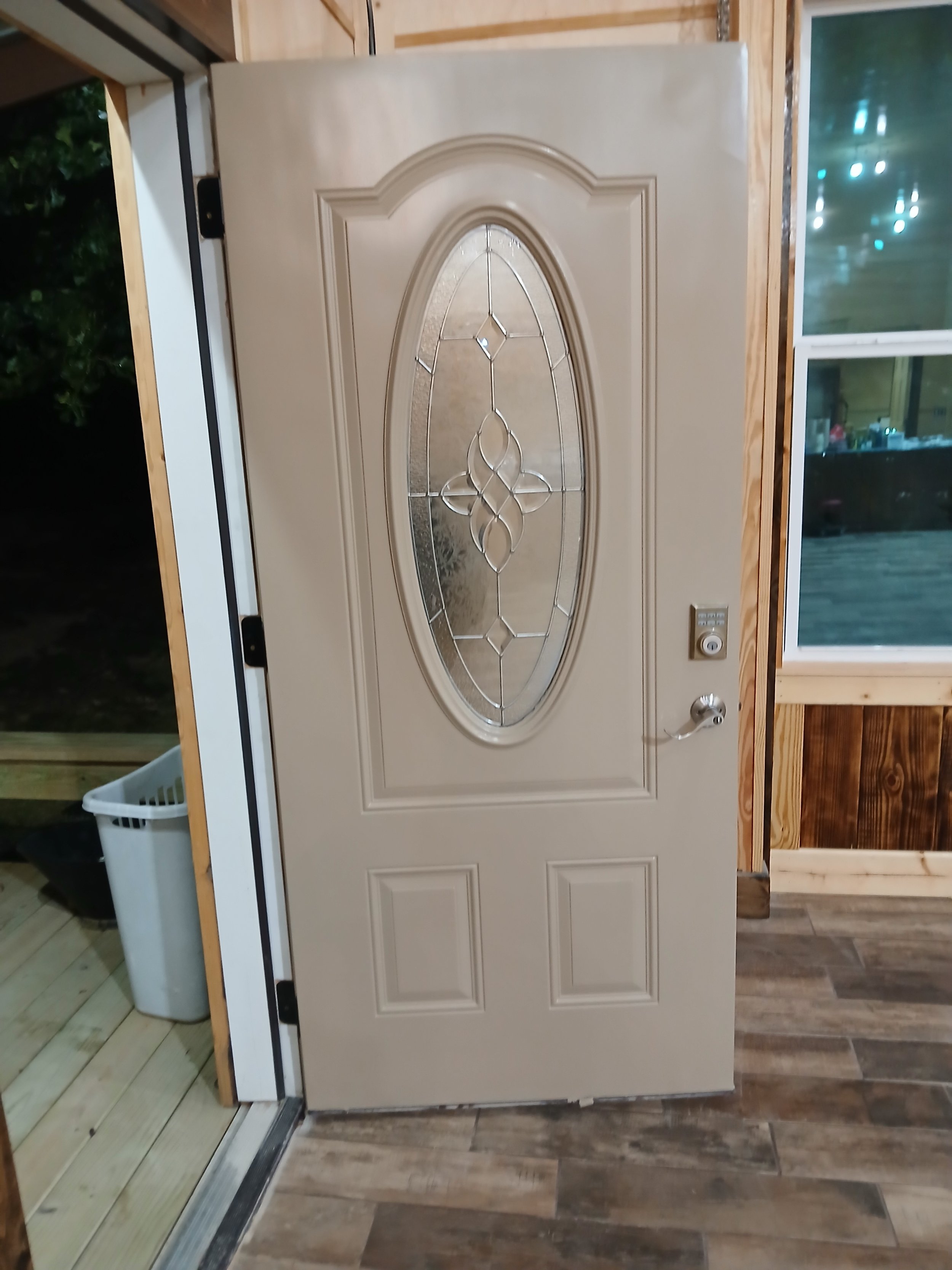 A beige decorative front door with an oval stained glass window and a metal handle, installed in a wooden-frame doorway. There is a doorbell or access control panel to the right of the door.