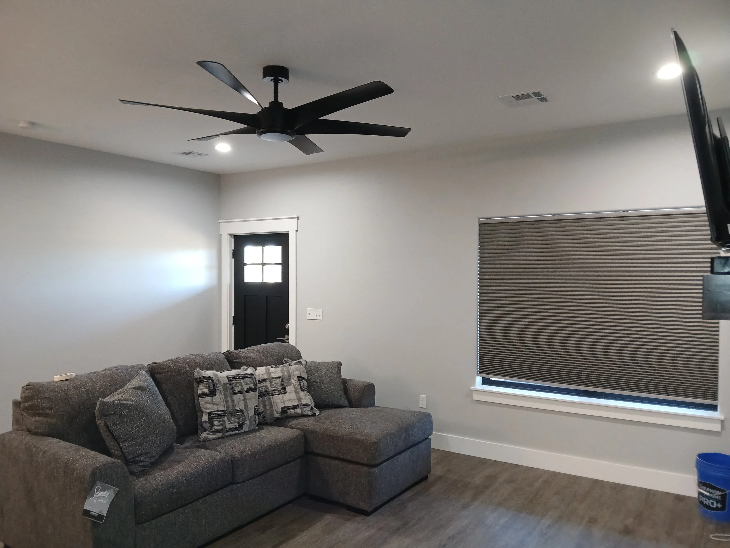 Living room with a gray sectional sofa with cushions, a window with closed blinds, a black front door with a small window, a ceiling fan, and a mounted TV on the right side.