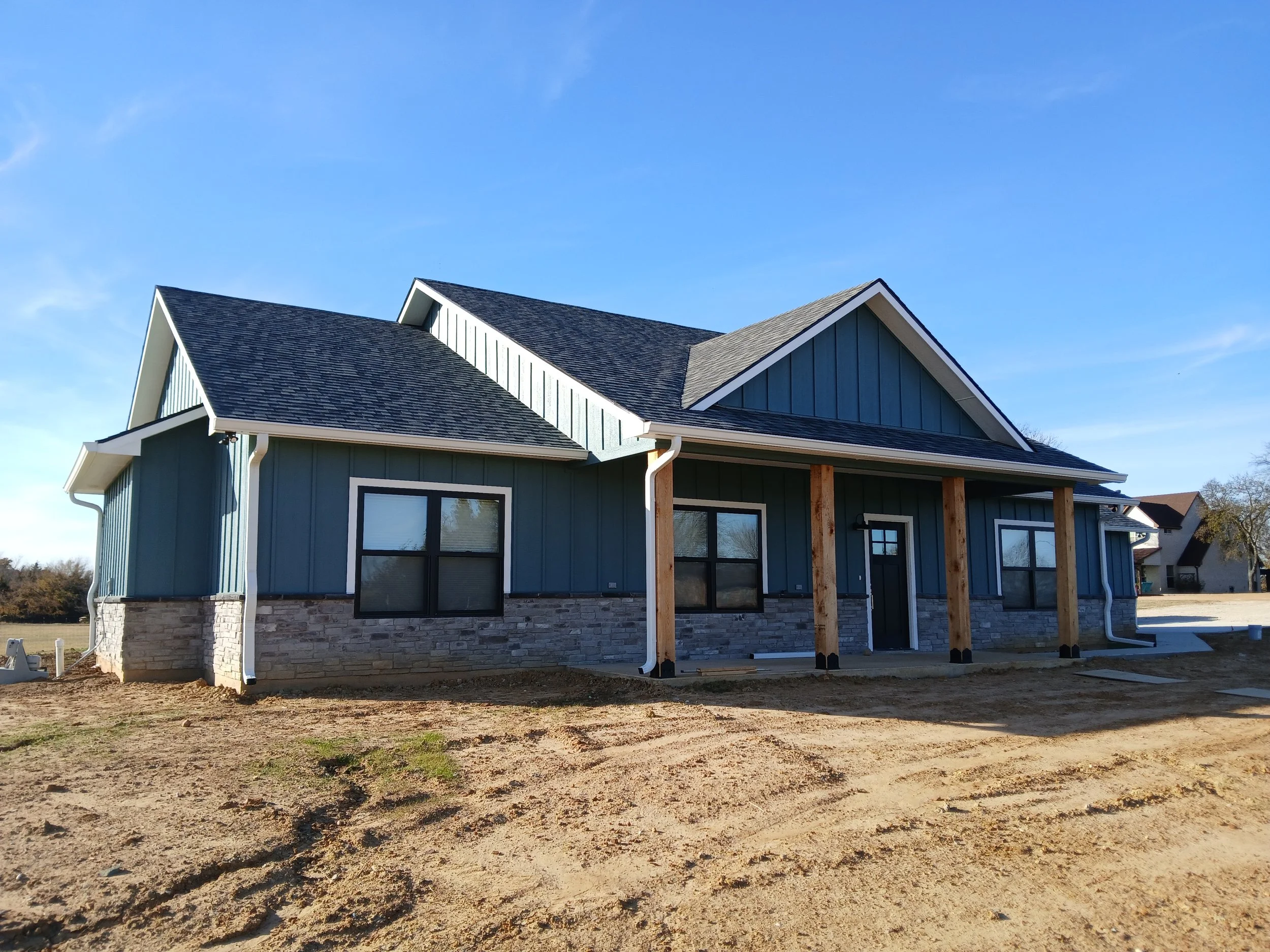 Newly constructed house with blue exterior, stone foundation, and wooden columns, under a blue sky, with construction ground in the foreground.