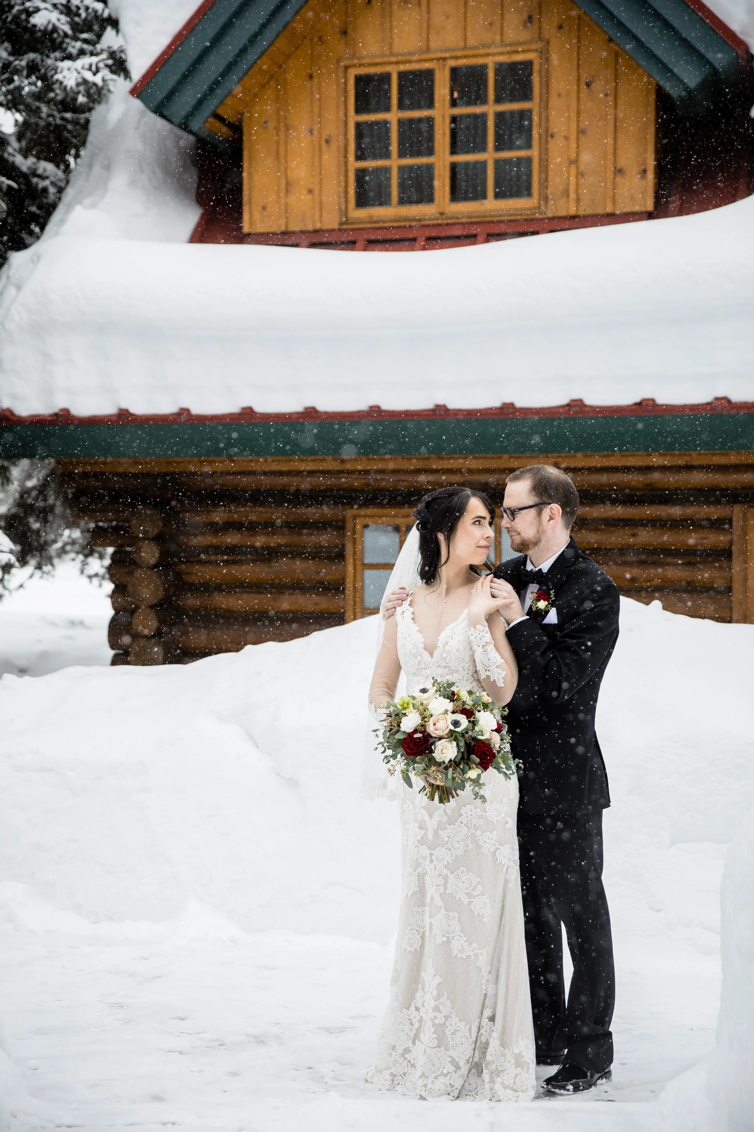 Bride and groom in wedding attire standing in snow outside a wooden cabin, with falling snowflakes.