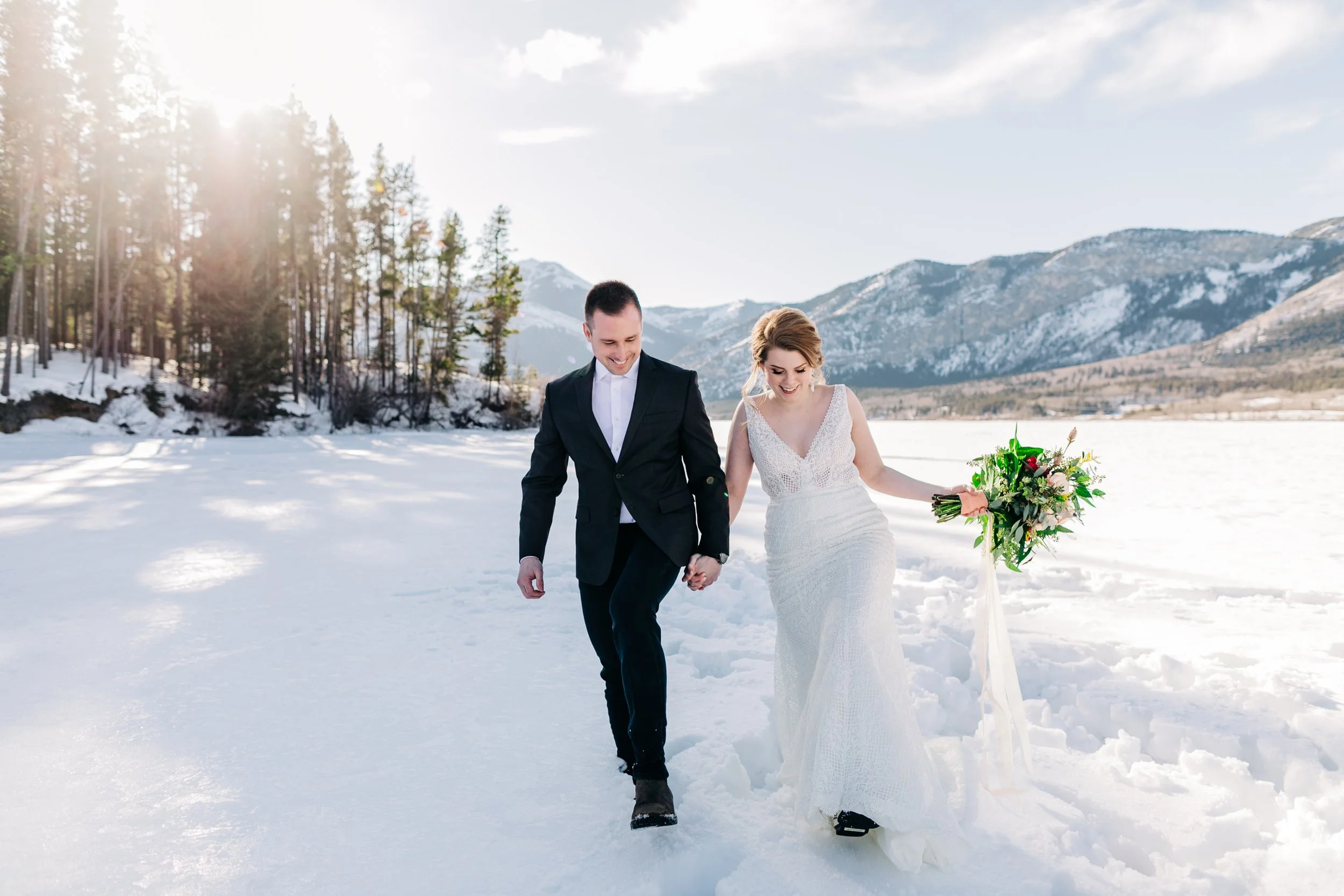 A bride and groom walking hand in hand through a snow-covered landscape with mountains and trees in the background, during daytime.