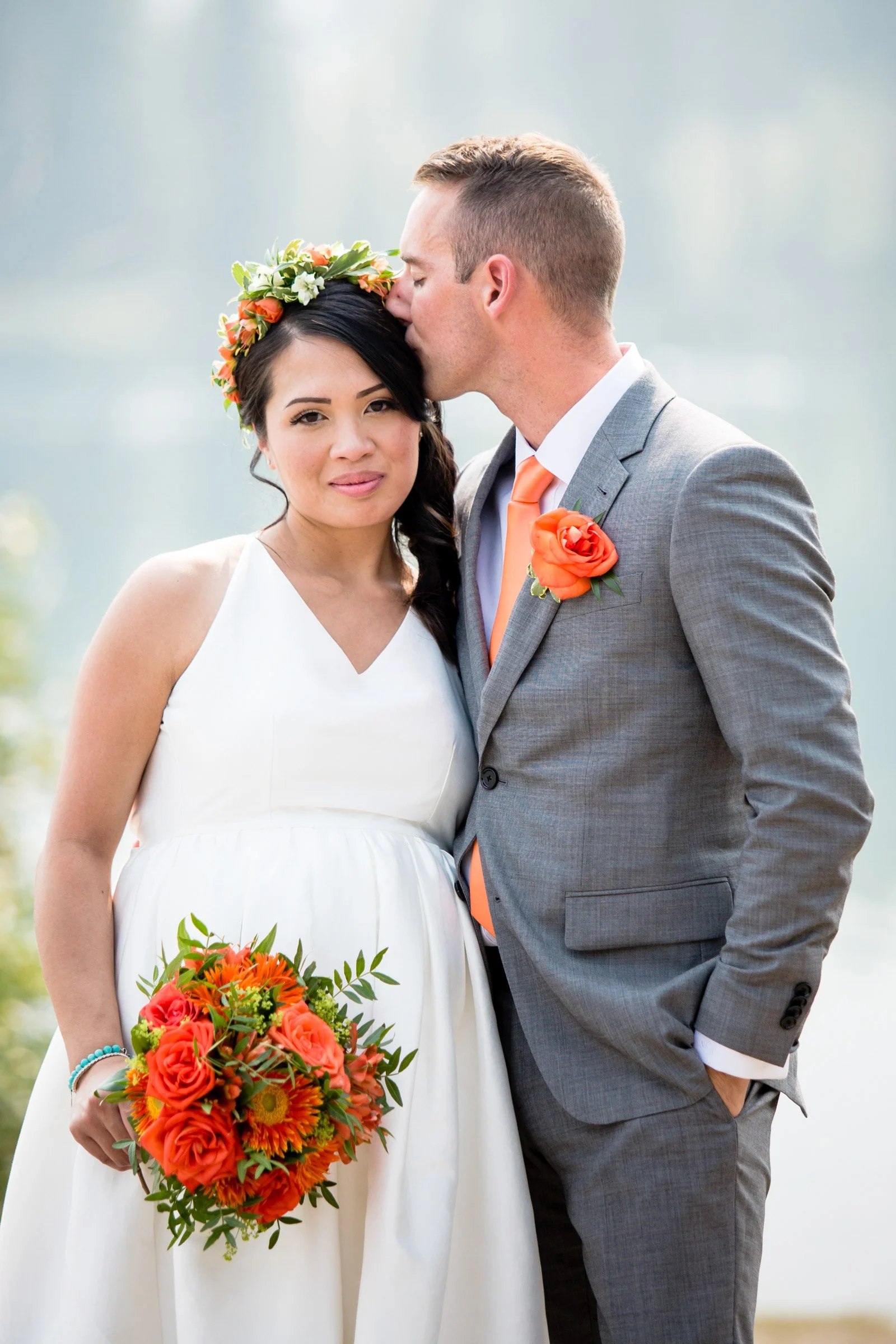 A newlywed couple; the bride in a white dress holding a colorful orange and pink bouquet, the groom in a gray suit with an orange tie and rose boutonniere, kissing her on the forehead.