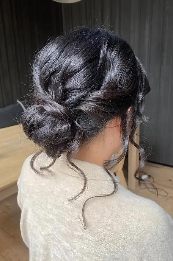 An elegant updo hairstyle with loose curls on a woman with dark hair, wearing a light-colored top, indoors with dark wooden paneling background.