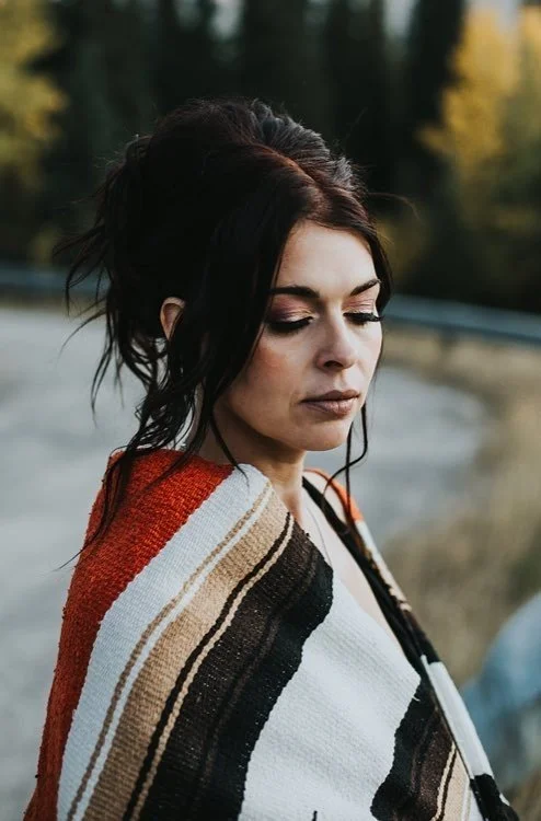 A woman with dark hair styled in an elegant updo, wearing a multicolored striped shawl, standing outdoors with a blurred background of trees and a road.