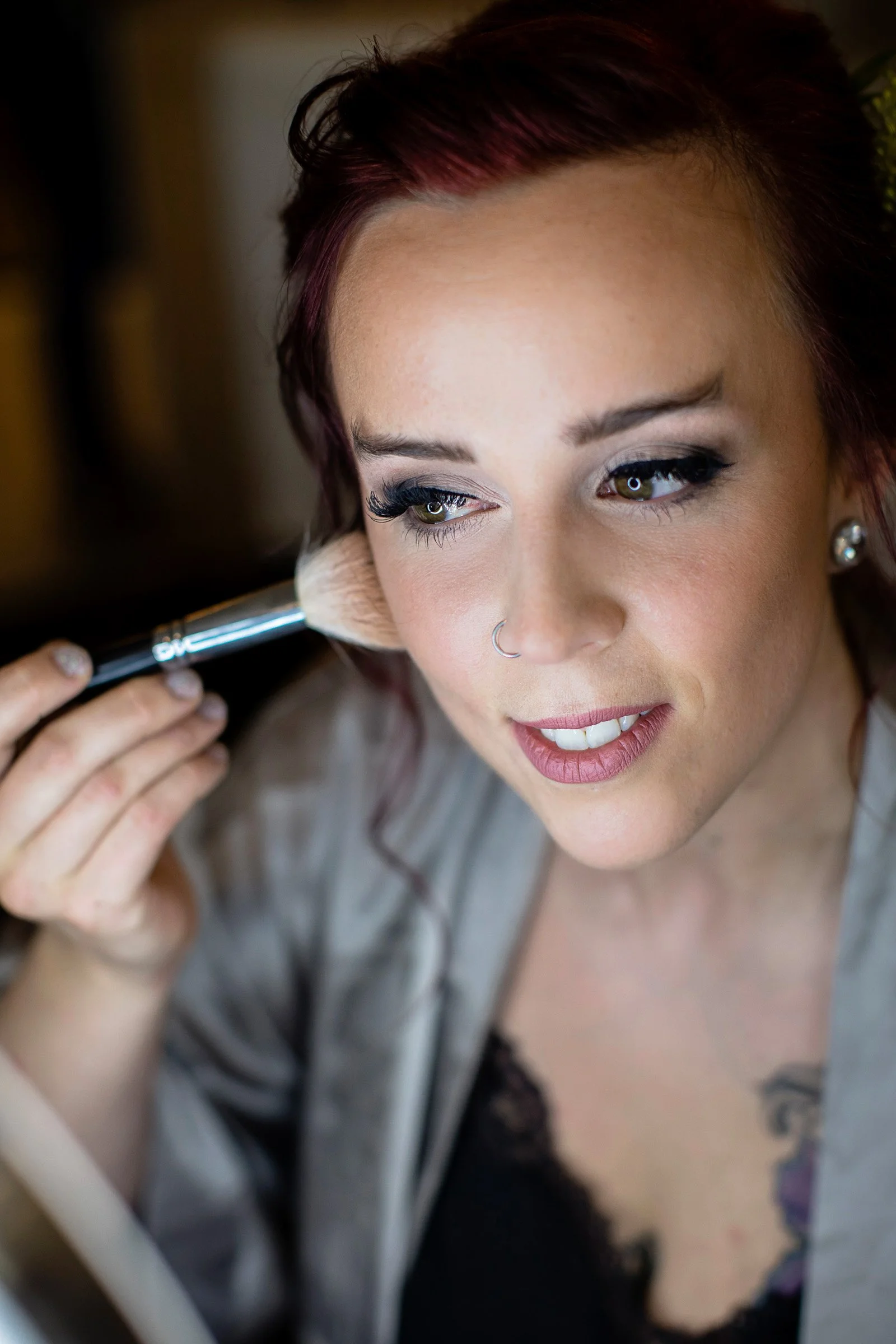 Woman with red hair having makeup application, using a large makeup brush on her cheek, wearing a gray satin robe.