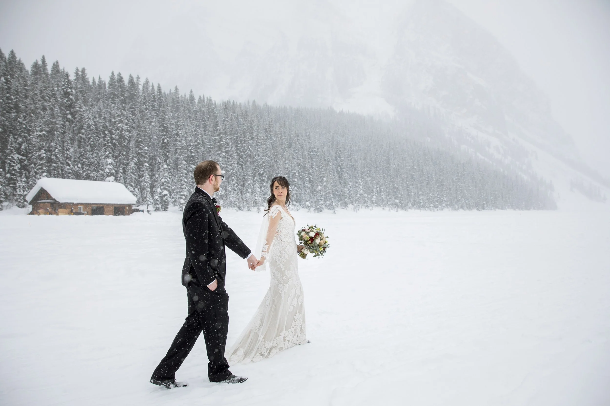 Bride and groom holding hands in a snowy landscape with a mountain and forest background