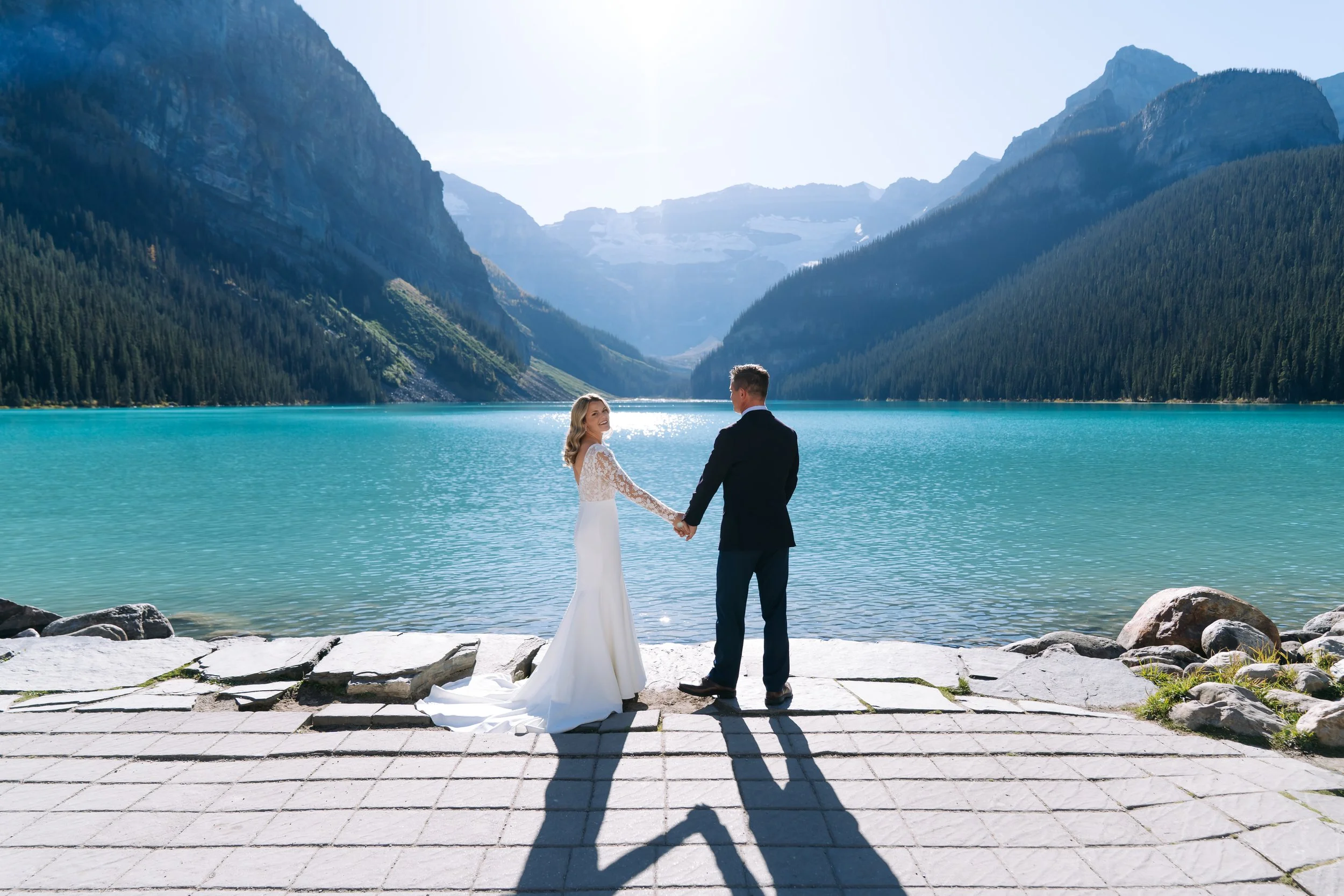 A bride and groom holding hands and facing each other by a lake with mountains in the background on a sunny day.