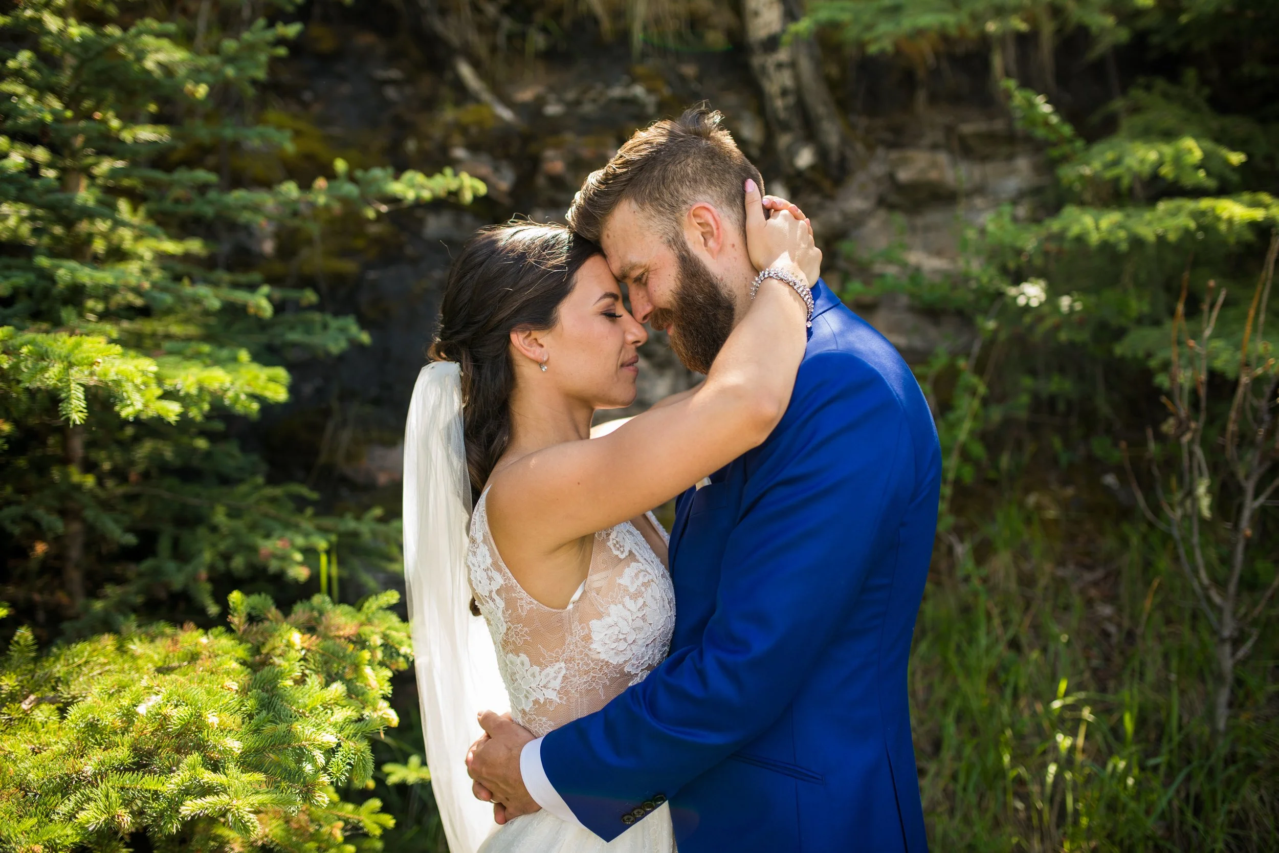 A bride and groom embracing outdoors with their foreheads touching, eyes closed, in a natural setting with greenery.
