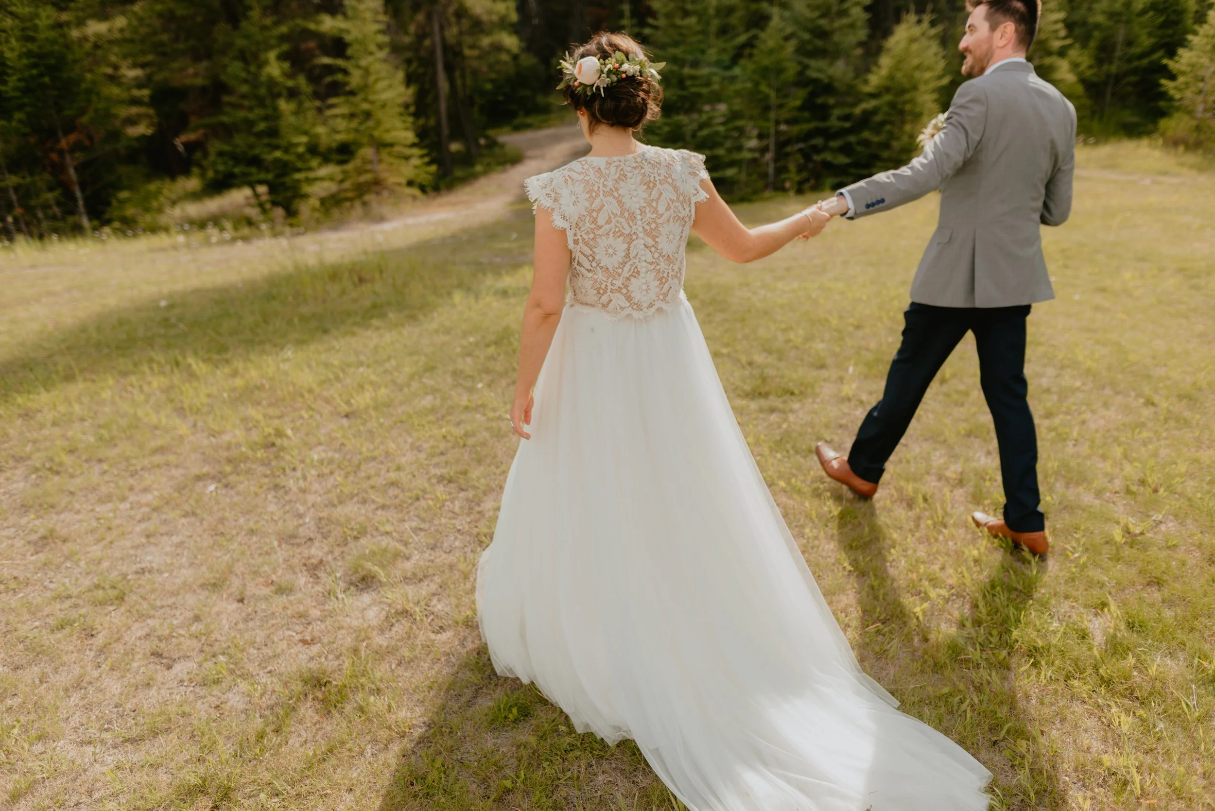 A bride and groom holding hands outdoors on a grassy field with trees in the background. The bride is wearing a white dress with lace details and a floral crown, and the groom is in a gray suit with dark pants and brown shoes.