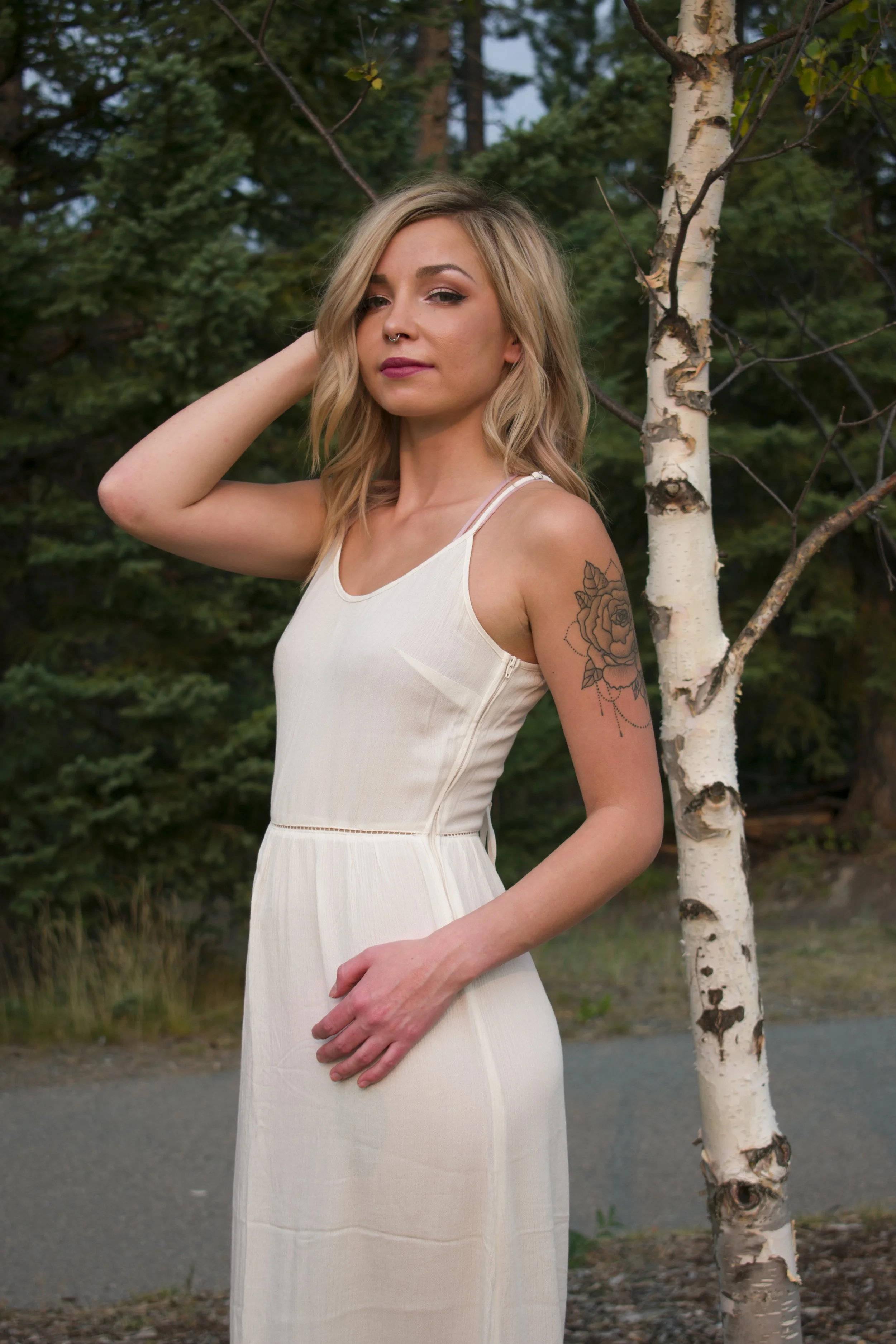 Young woman in a white dress standing outdoors near a birch tree, with a background of green trees.
