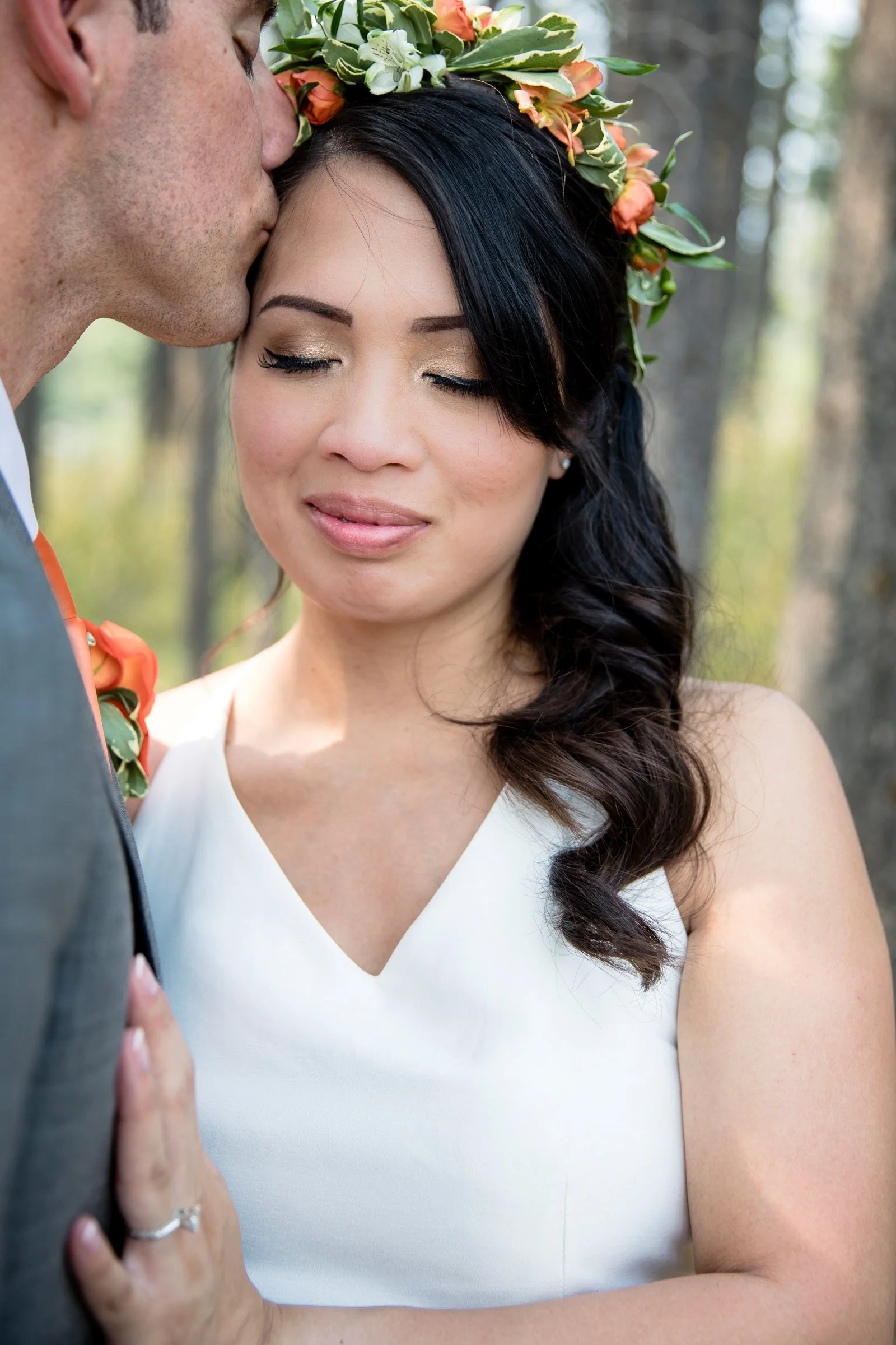 A bride with closed eyes and a floral crown gently presses her hand to a groom's chest as he kisses her forehead, standing outdoors with trees in the background.