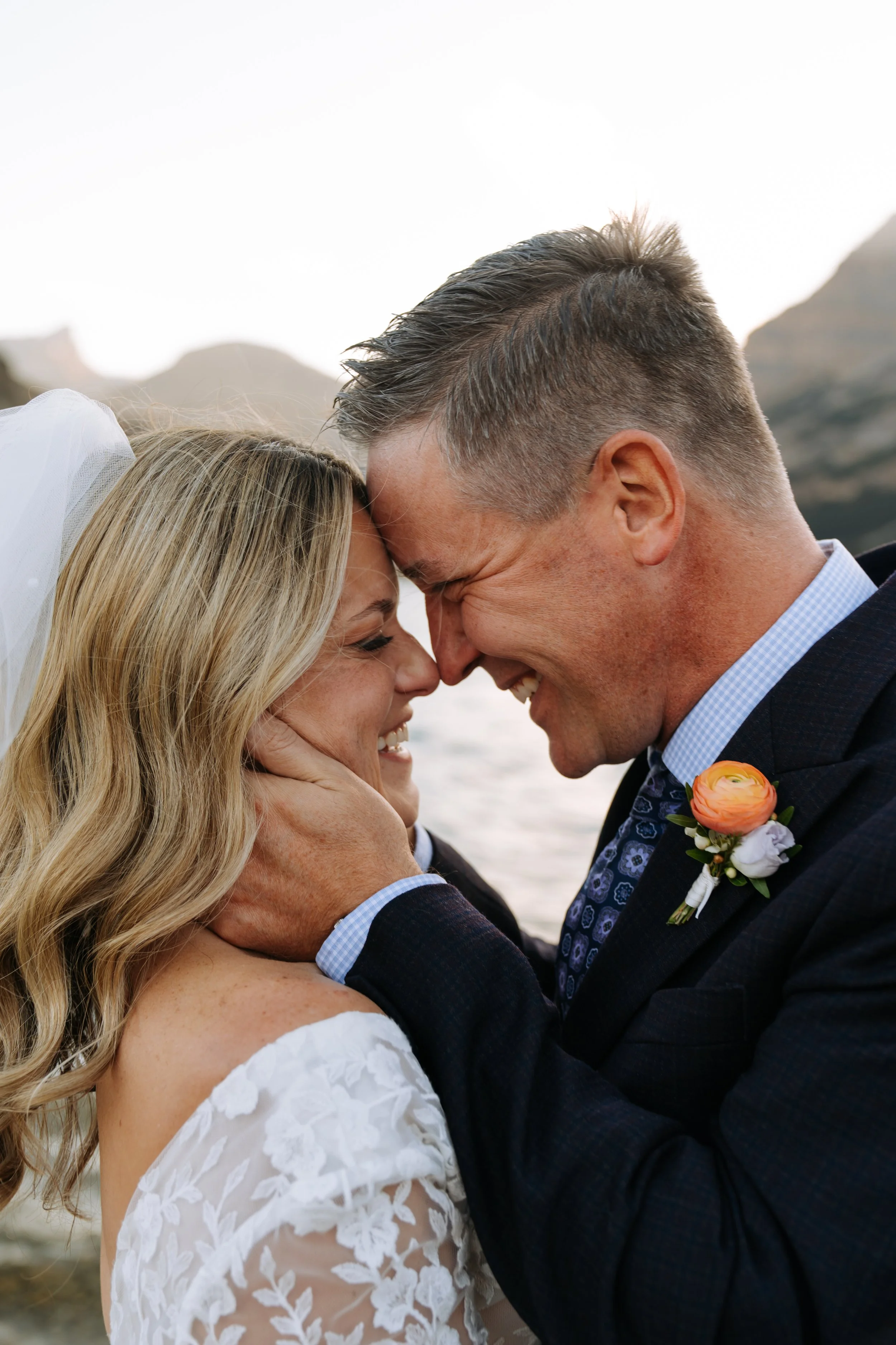 A couple on their wedding day, smiling and touching foreheads, with outdoor mountains in the background.