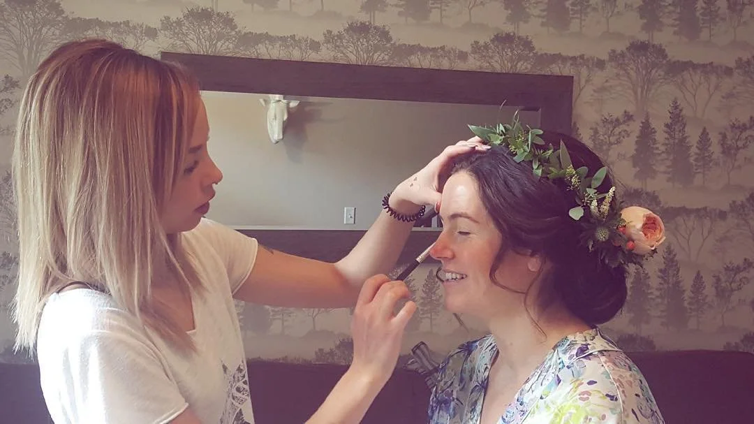A woman with dark hair wearing a floral crown is having her makeup done by a makeup artist.
