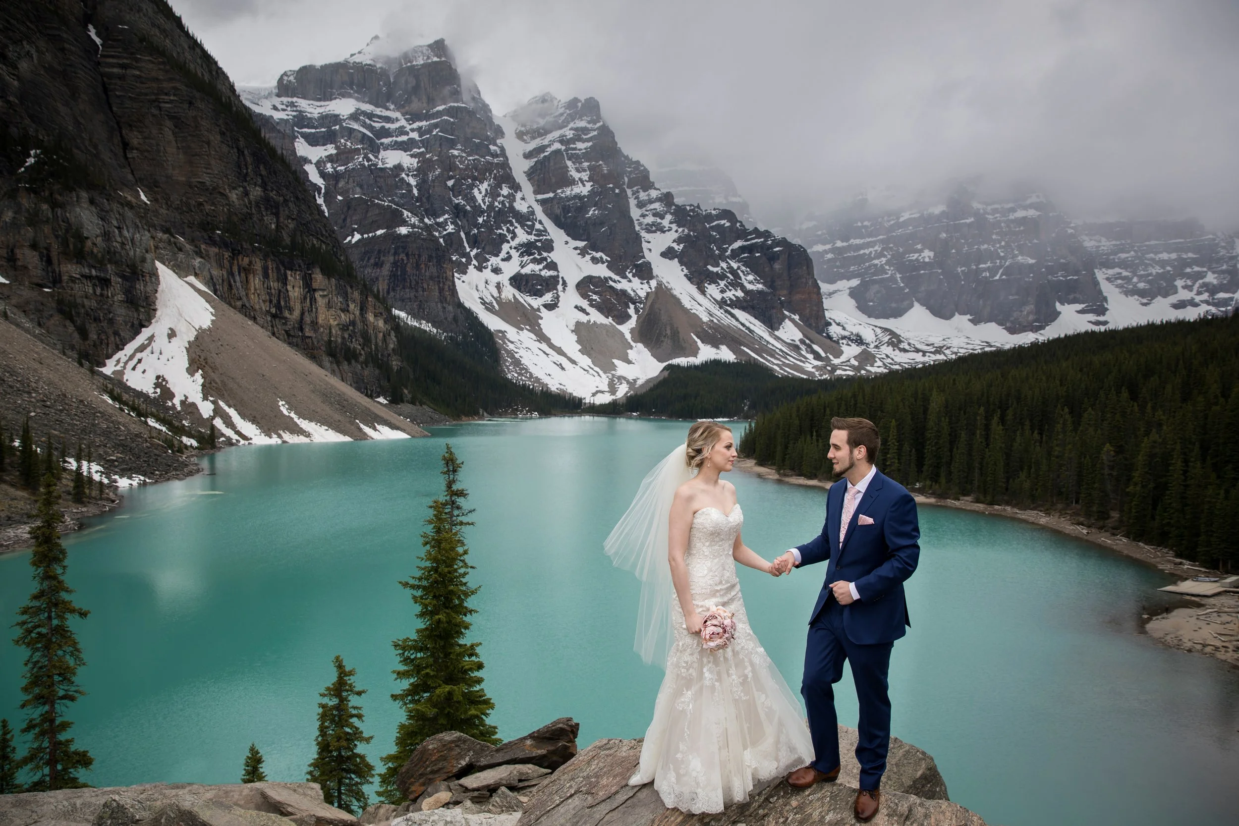 A bride and groom hold hands on a rocky ledge overlooking a turquoise lake with forested mountains in the background.