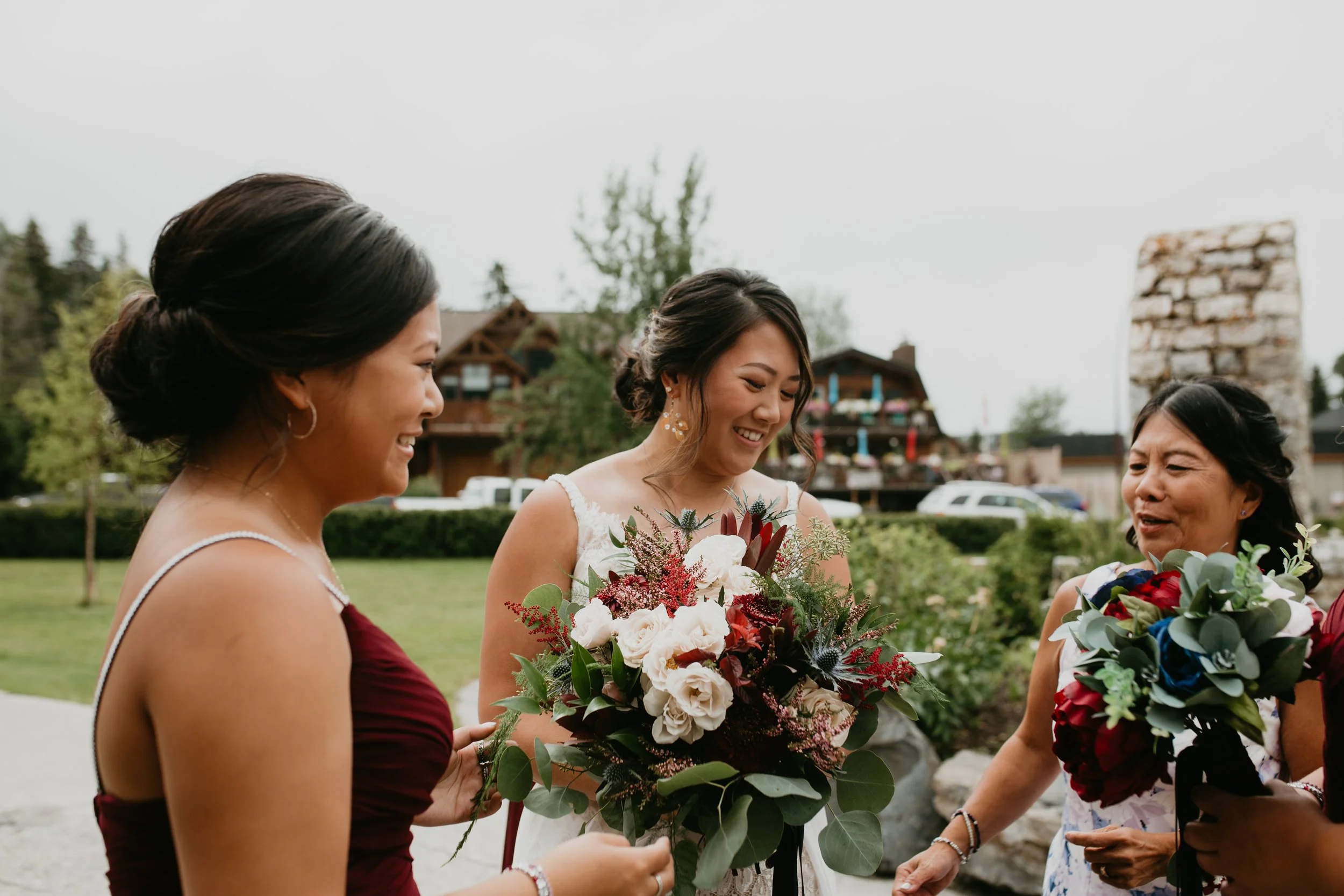 Three women smiling and chatting outdoors, two are dressed in burgundy and white dresses holding bouquets, with one woman in a white wedding gown. The background shows a grassy area, trees, and residential buildings.