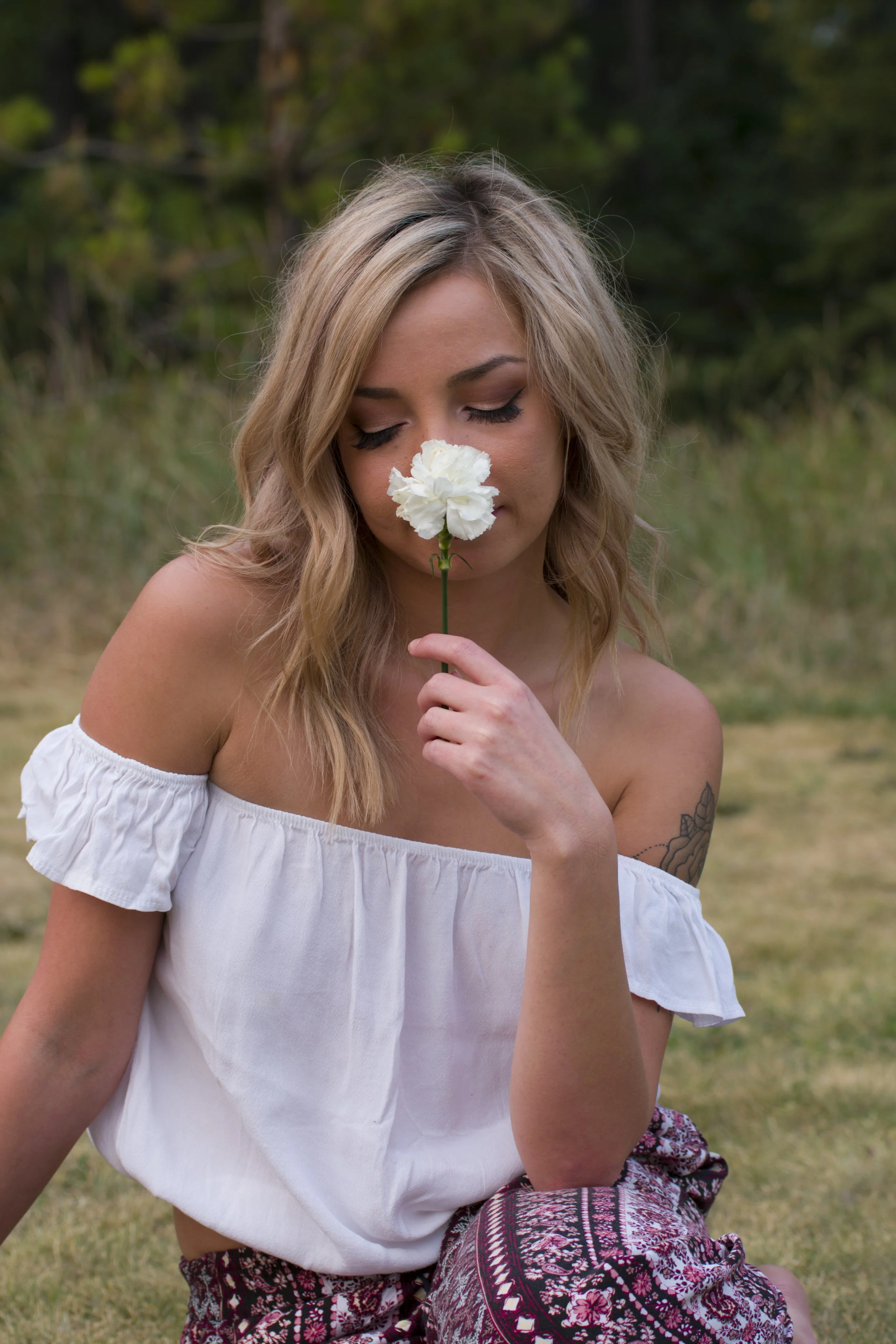 A young woman with blonde hair, wearing an off-the-shoulder white top and patterned pants, sitting outdoors on the grass, holding a white flower near her nose with her eyes closed.