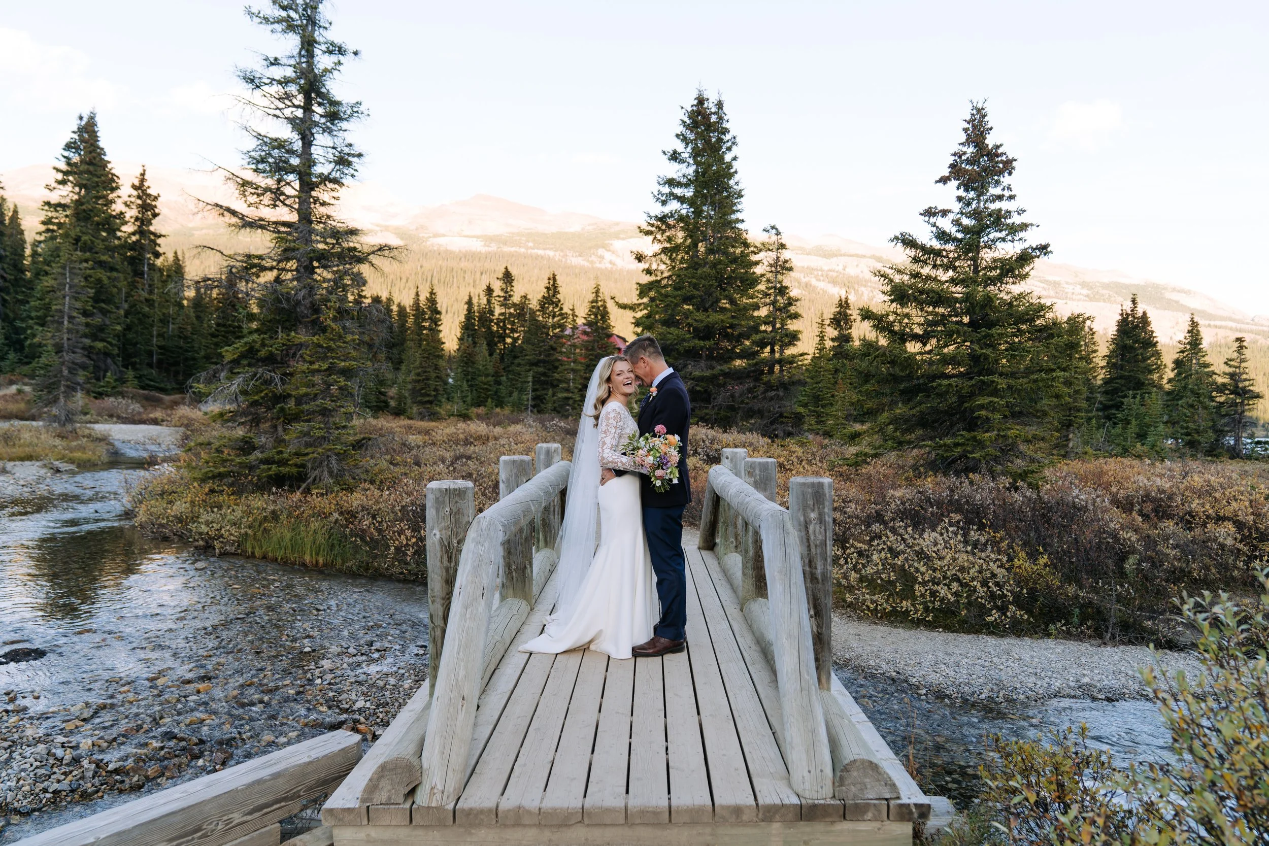 A newlywed couple standing on a wooden bridge over a river, embracing and smiling in a mountainous, forested landscape during daylight.