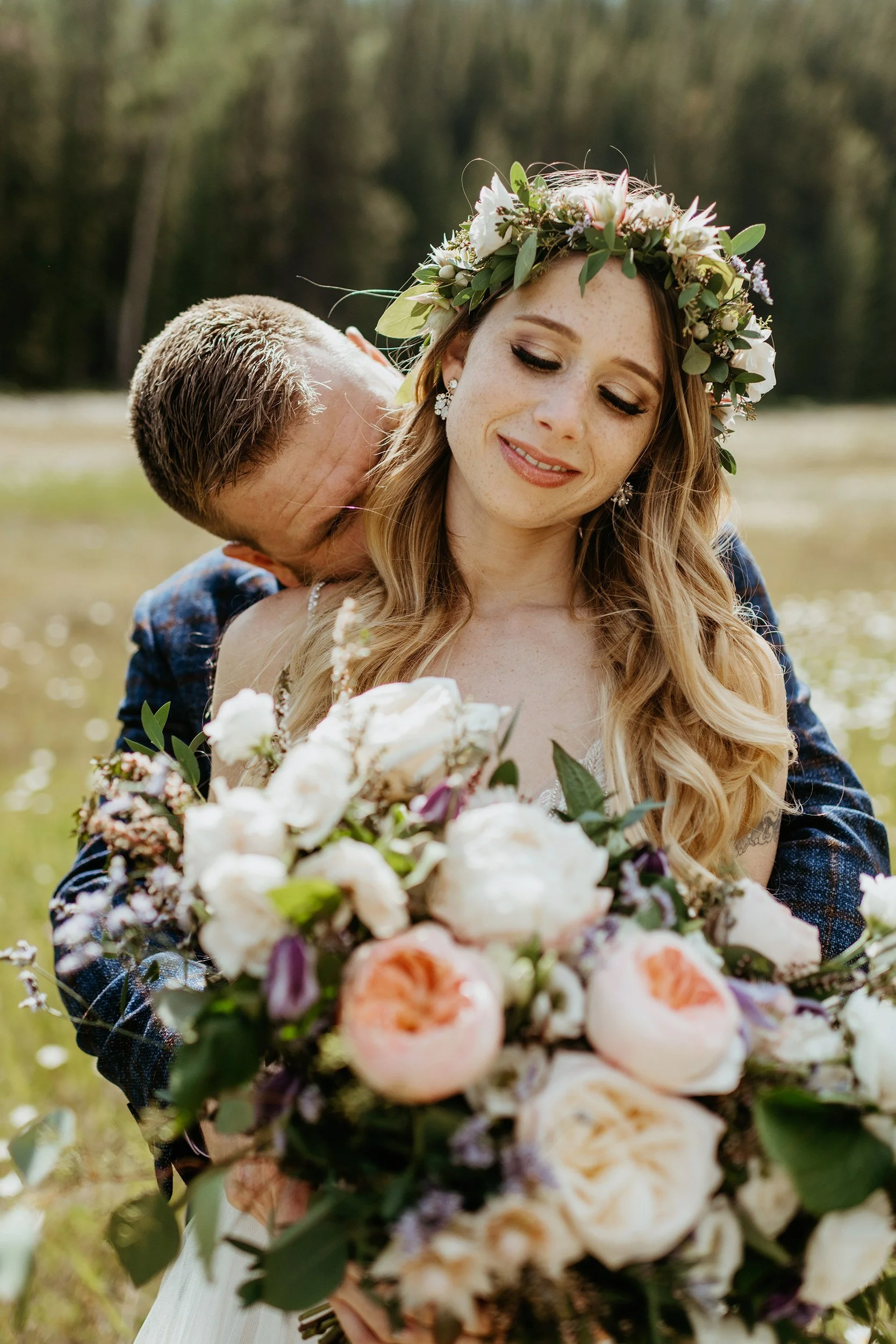 A bride with a floral crown holding a large bouquet of flowers, with a man kissing her neck outdoors.