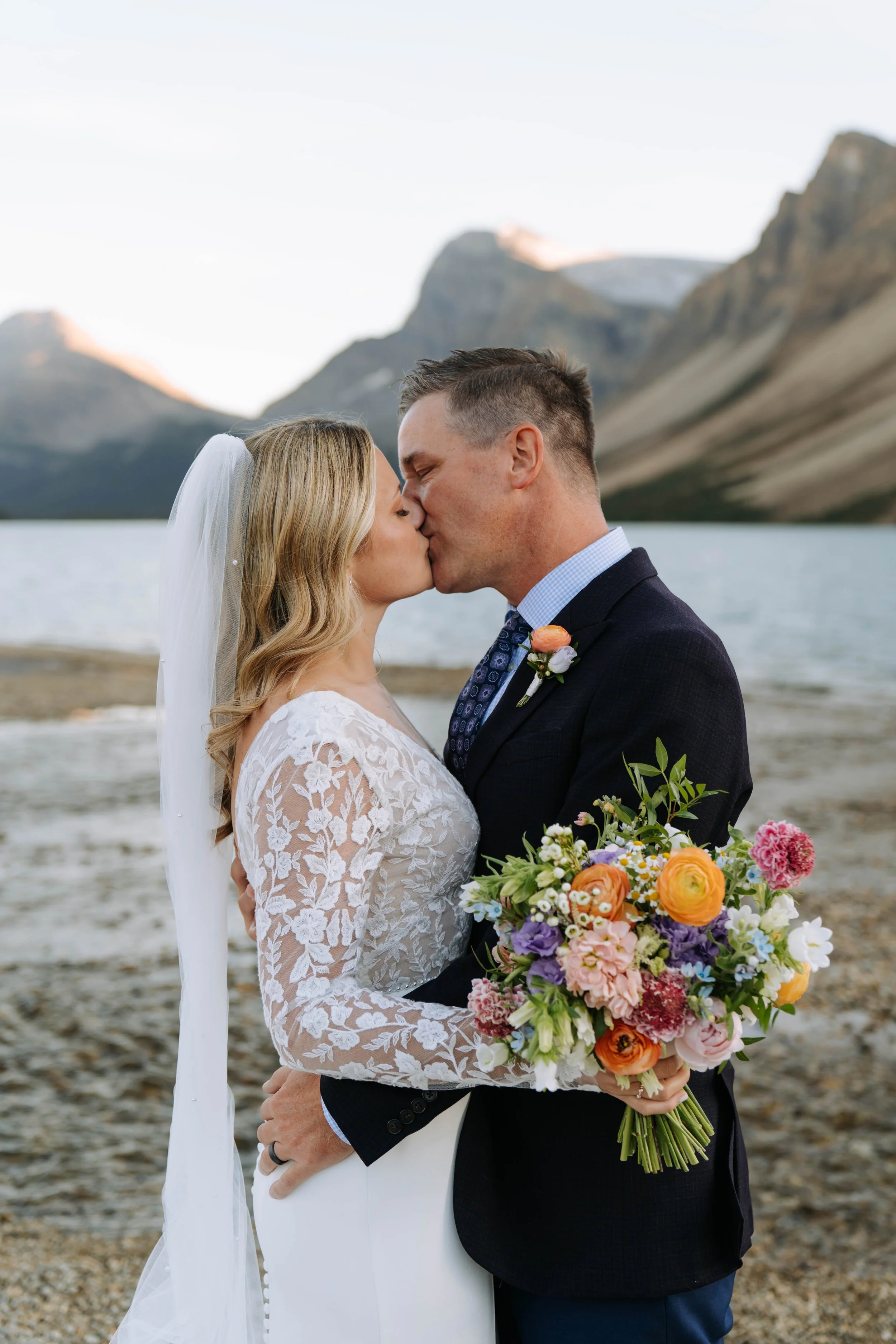 A newlywed couple sharing a kiss by a lake with mountains in the background. The bride holds a colorful bouquet of flowers, and the groom wears a dark suit with a boutonniere.