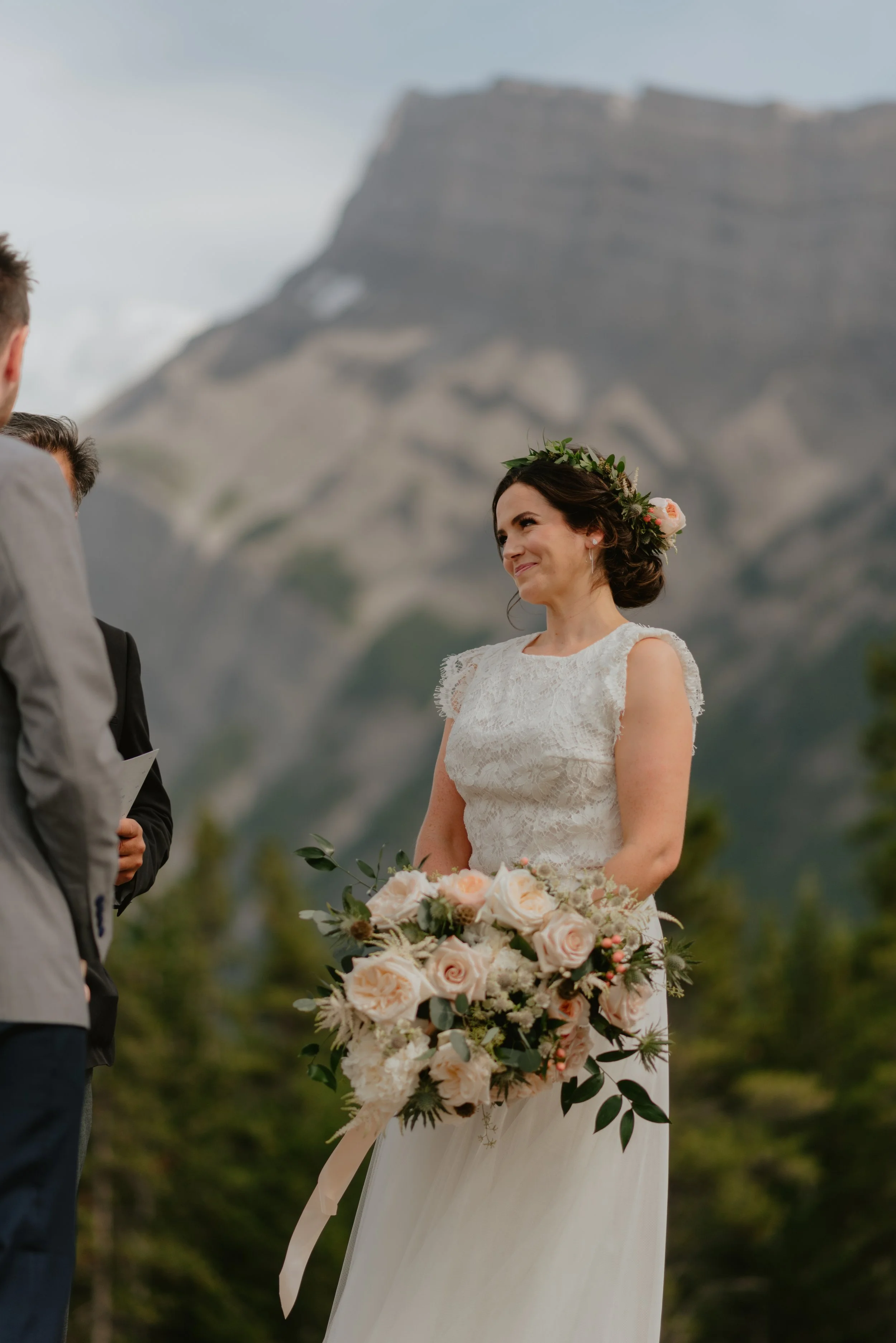 Bride holding a large bouquet of pale pink roses, white flowers, and greenery during an outdoor wedding ceremony with mountains in the background.