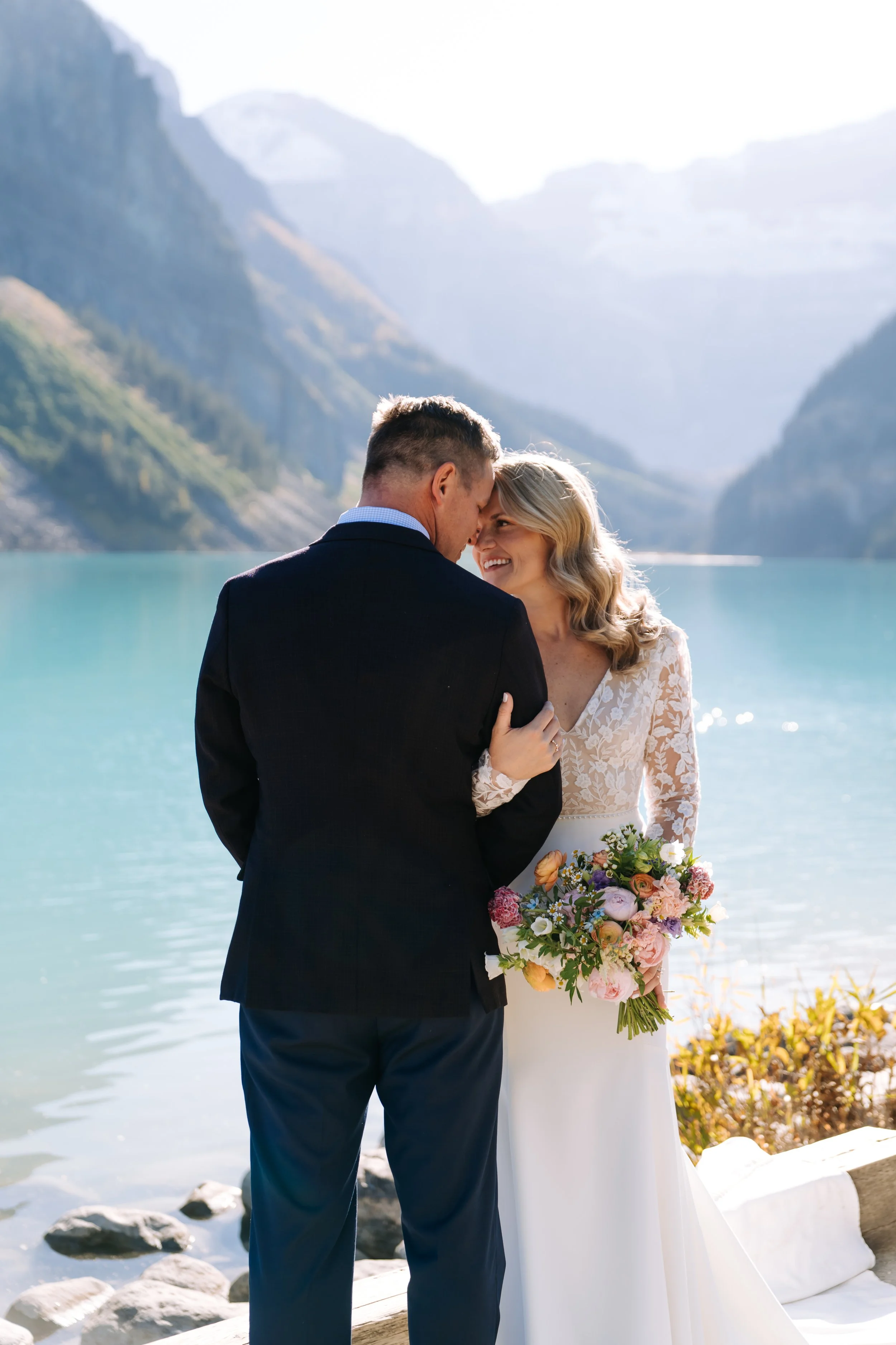 A bride and groom in wedding attire embrace by a lake surrounded by mountains, with the bride holding a bouquet of flowers.