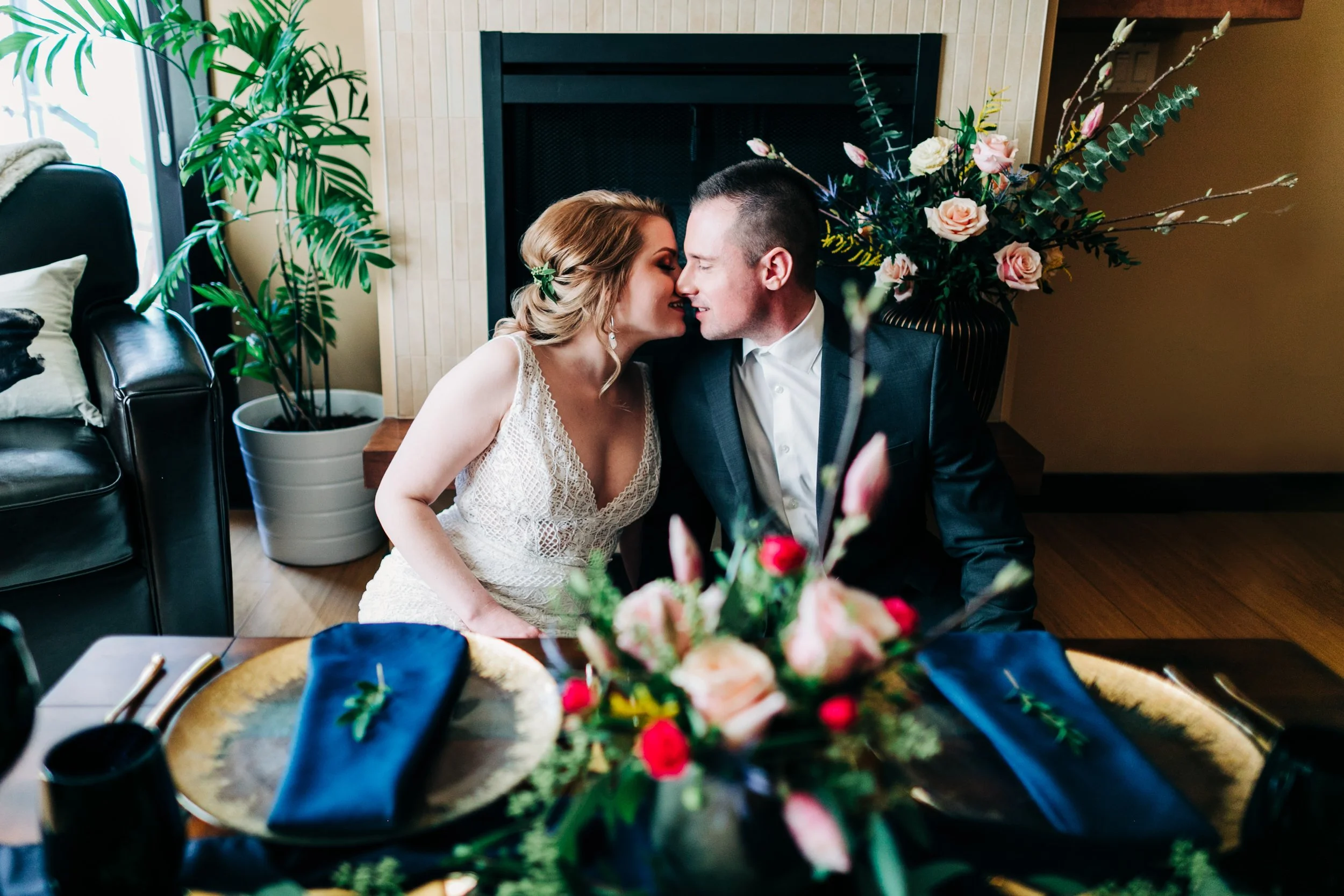 A bride and groom are close together, touching noses, in a warmly decorated living room with a fireplace and a floral arrangement in the background.