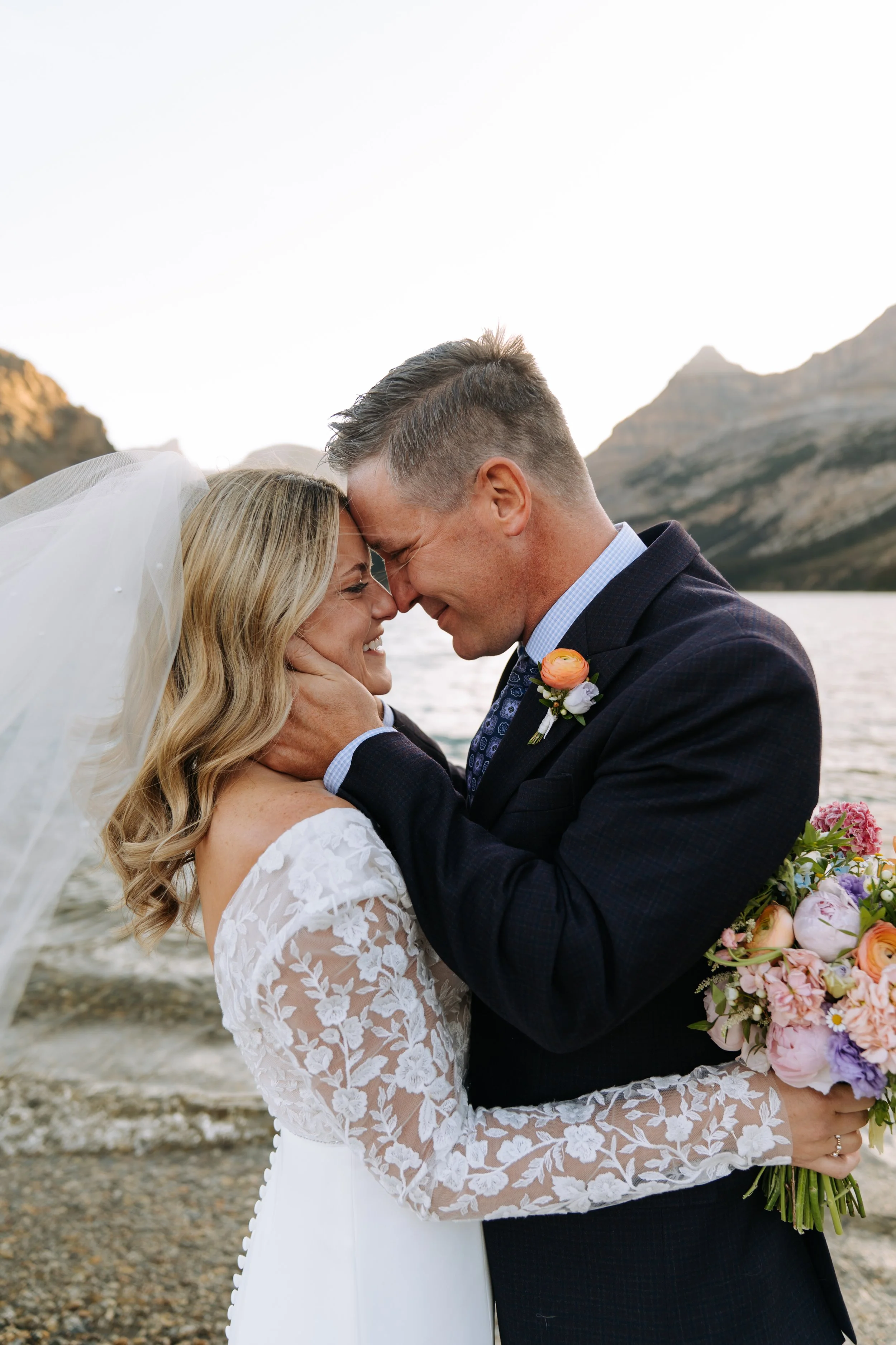 A bride and groom are outdoors on a rocky shore at sunset, embracing each other with their foreheads touching and smiling. The bride is holding a bouquet of flowers and wearing a white wedding dress with lace sleeves. The groom is wearing a dark suit
