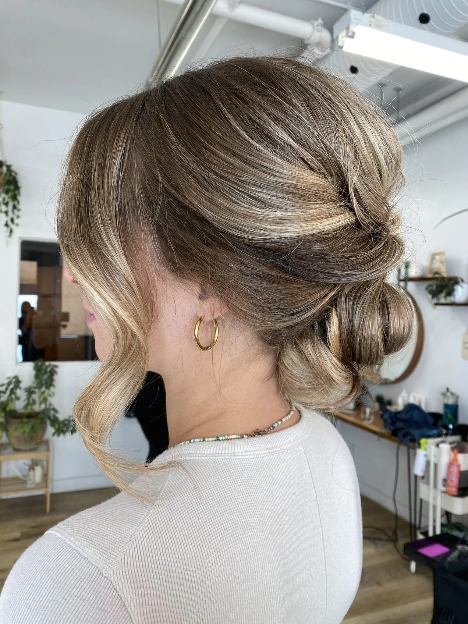 Woman with styled blonde hair in a bun, wearing gold hoop earrings and a beaded necklace inside a salon.