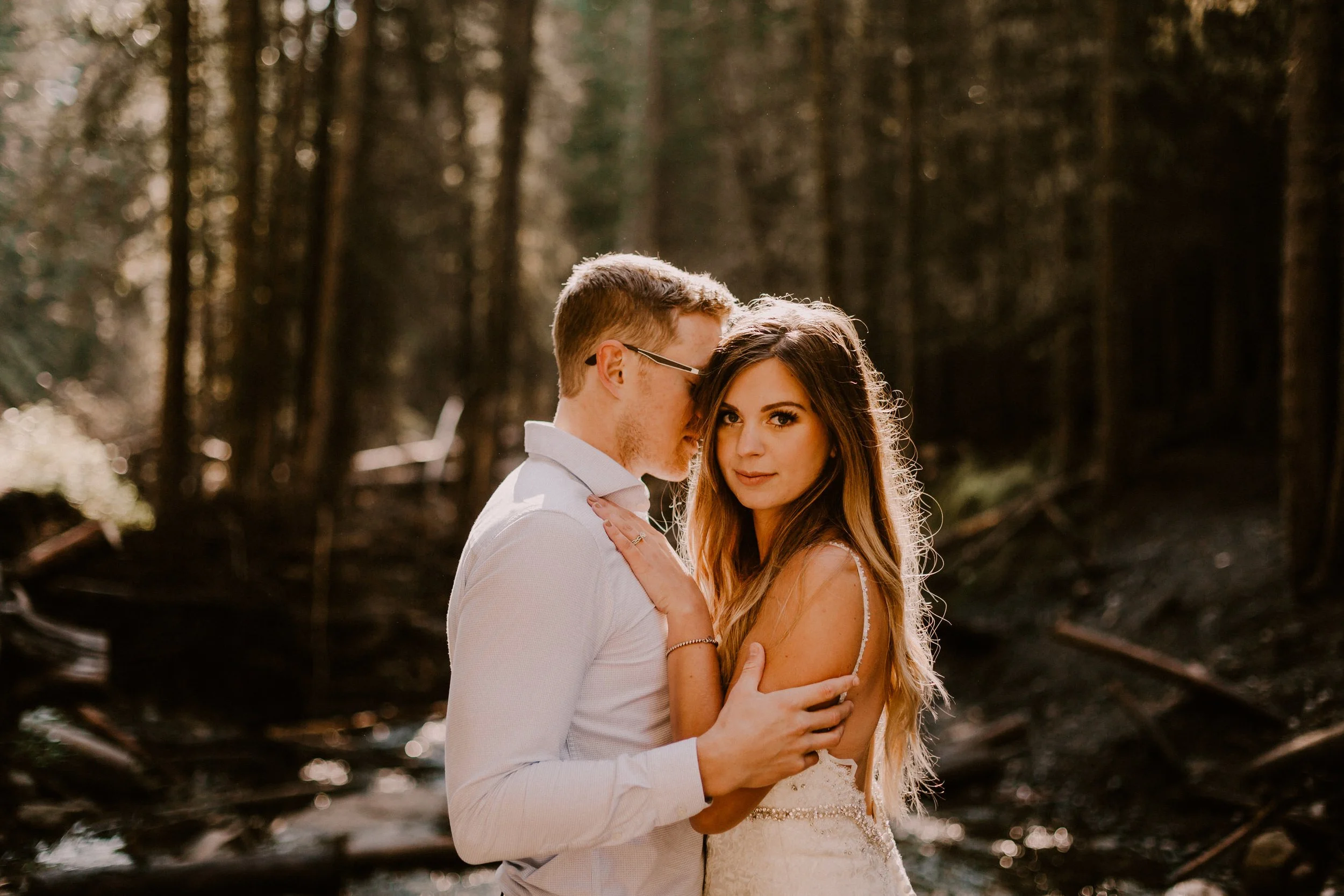 A couple in wedding attire standing close together in a forest, the man whispering into the woman's ear, with trees and sunlight in the background.