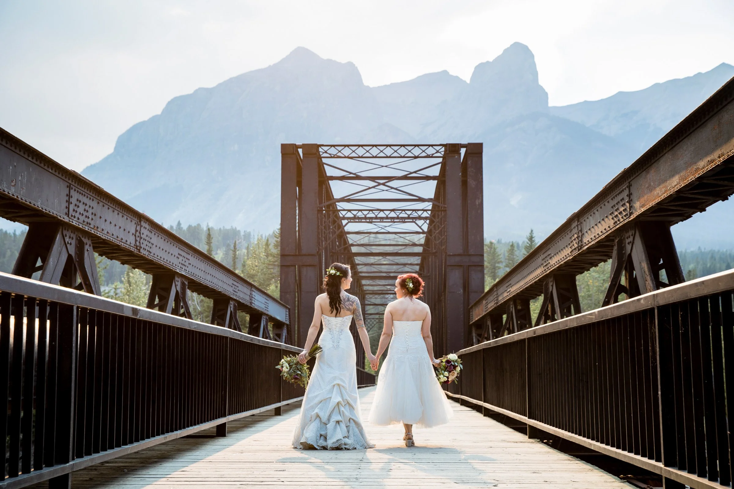 Two women in wedding dresses holding hands, walking on a wooden bridge with mountain scenery in the background.