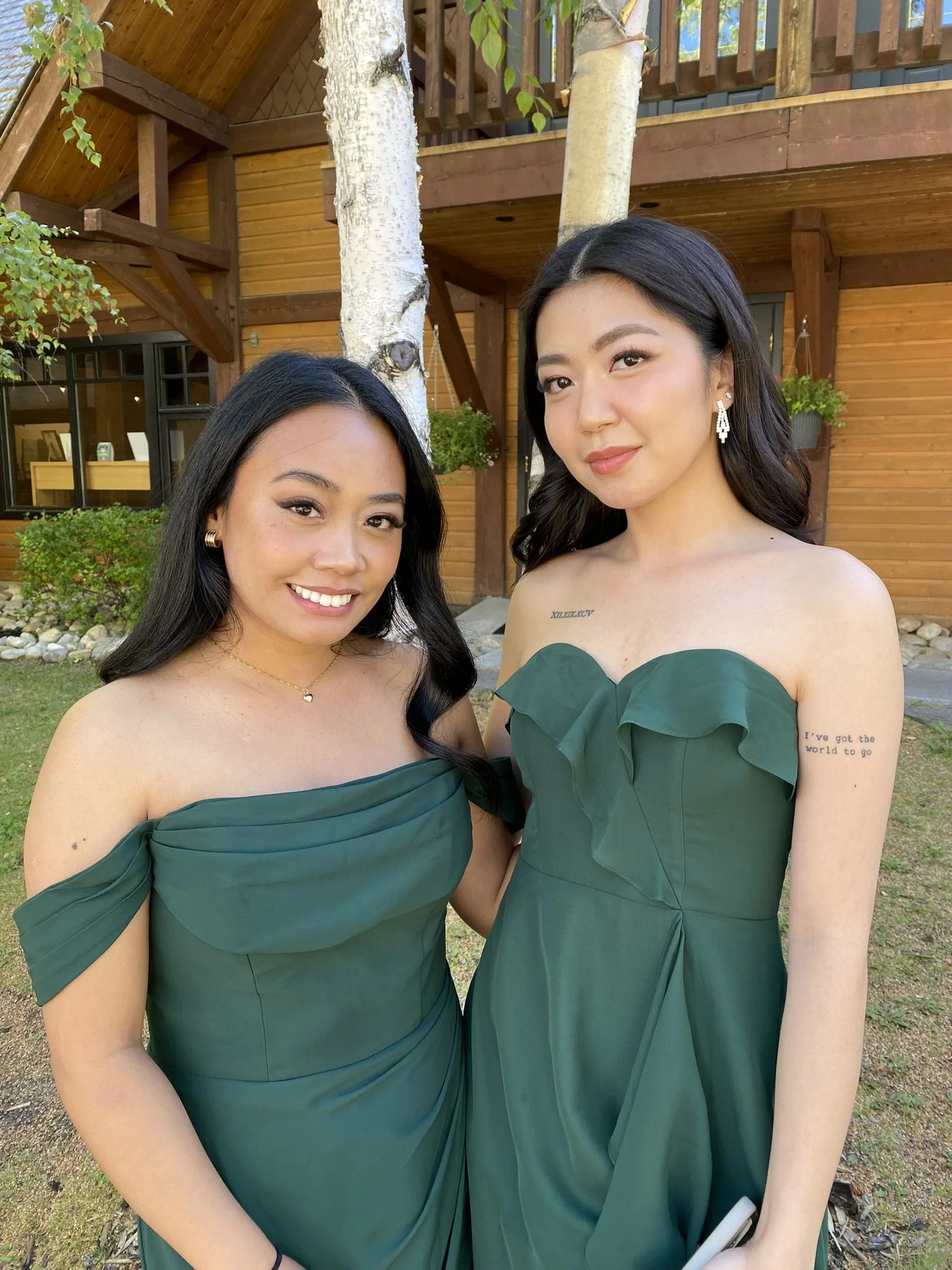 Two young women dressed in green strapless dresses standing outdoors, smiling and posing for a photo in front of a wooden house with trees and bushes.