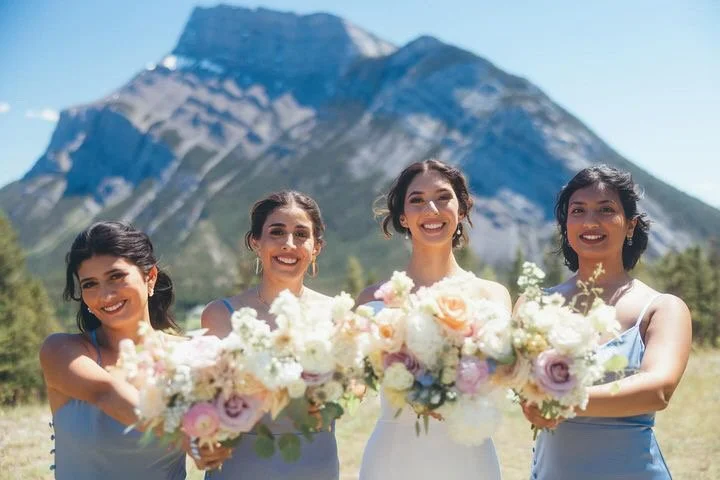 Four women in light blue and white dresses holding bouquets of flowers with a mountain in the background.