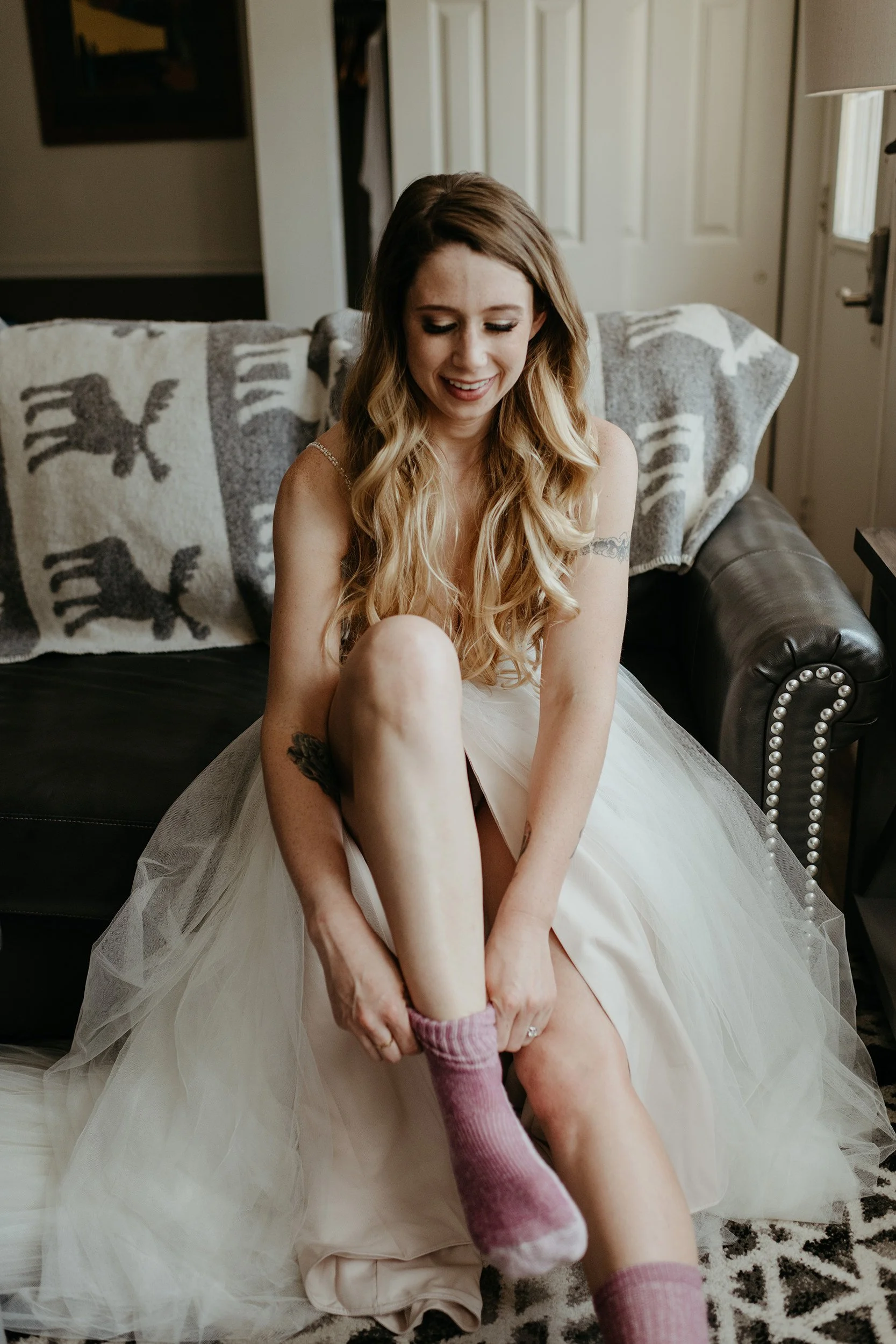 A woman in a white dress sitting on a couch, putting on pink fuzzy socks.