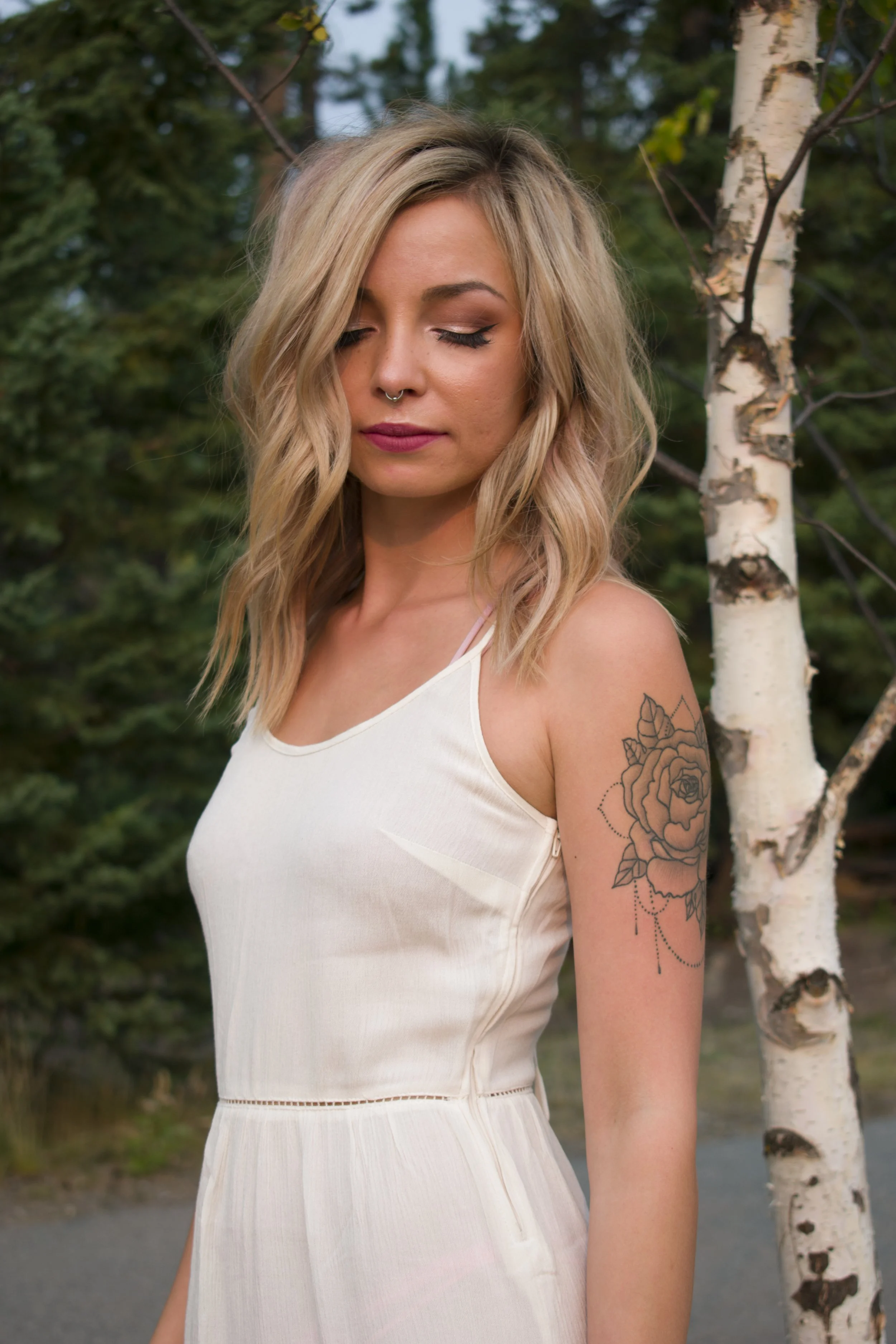 A young woman with blonde wavy hair standing outdoors near a white birch tree, wearing a white sleeveless dress, with her eyes closed and a tattoo of a rose on her upper arm.