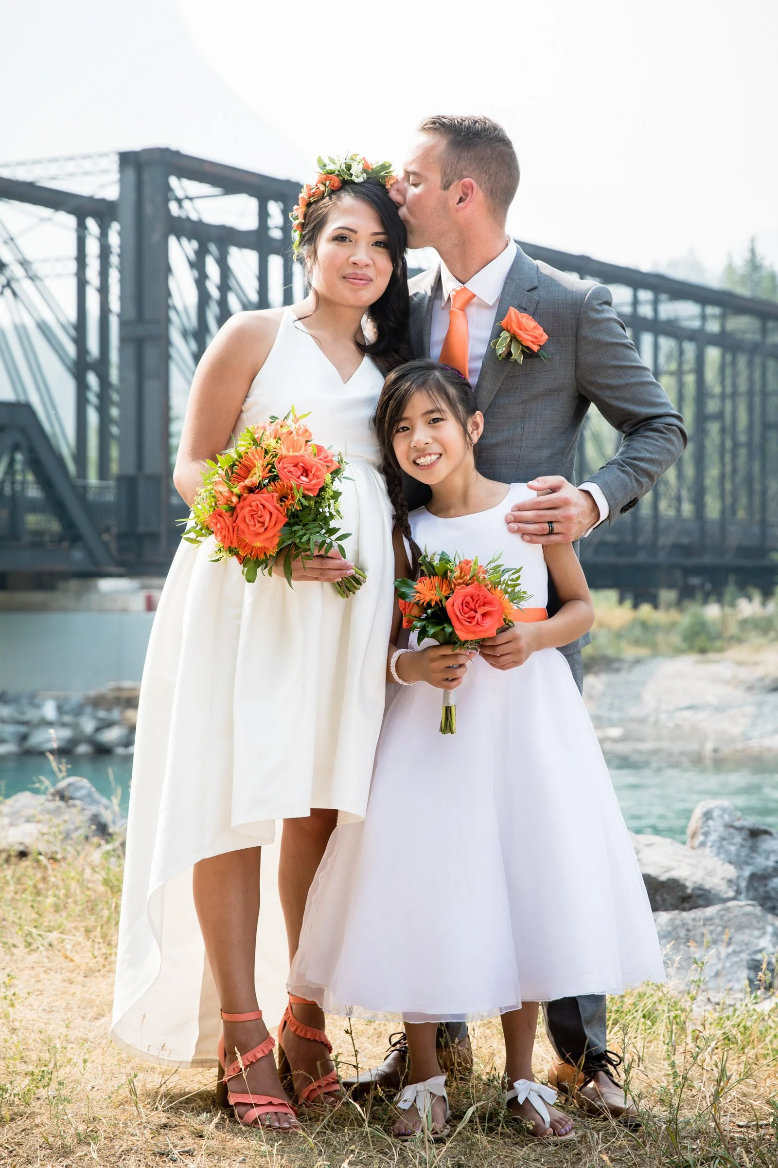 A bride and groom with a young girl, all holding orange and pink bouquets, standing outdoors near a river with a bridge in the background. The groom is kissing the bride on the forehead, and the girl is smiling at the camera.