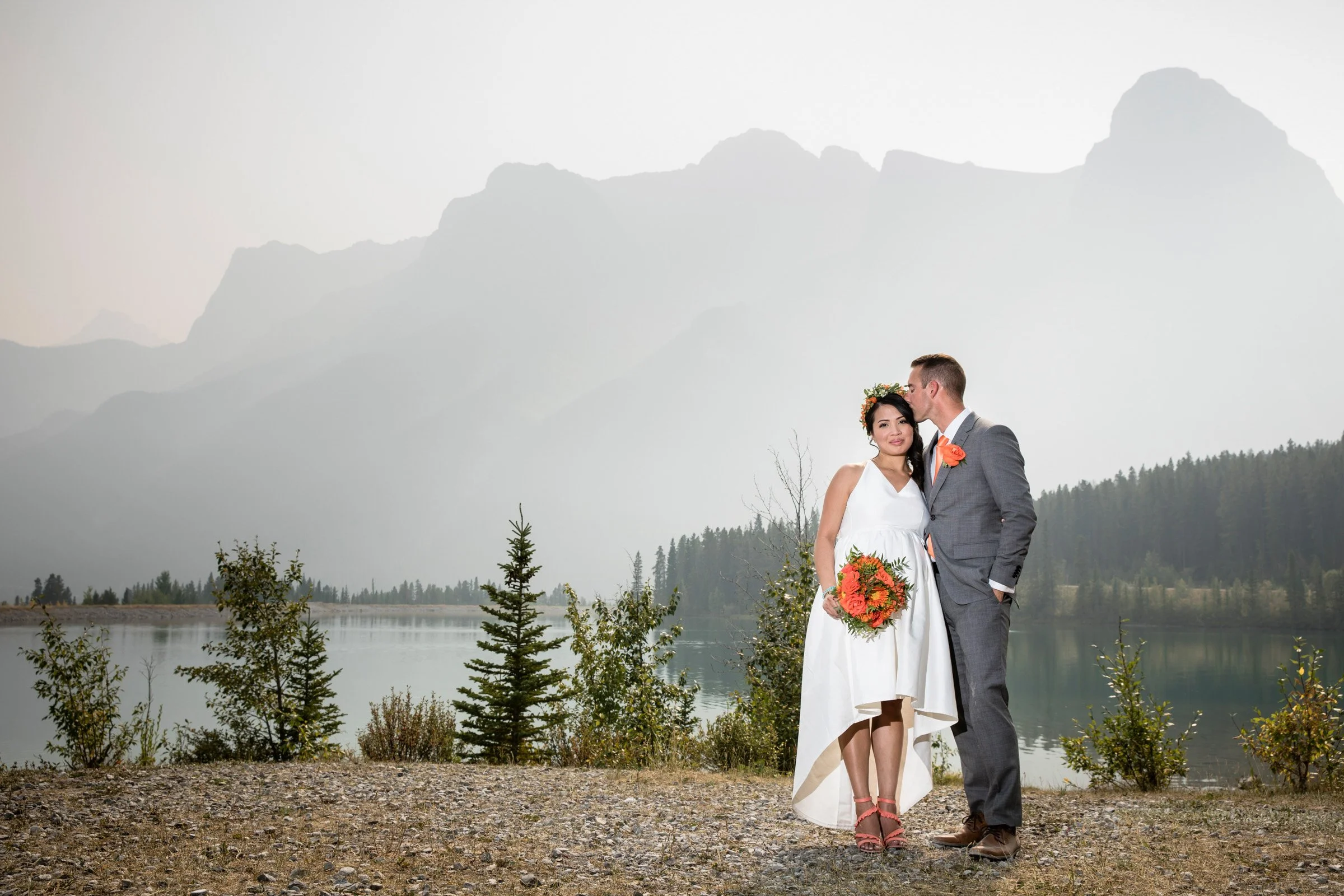 A wedding couple stands by a lake with mountains in the background. The bride is in a white dress holding a bouquet of flowers, and the groom is in a gray suit with a boutonniere, kissing her on the cheek.