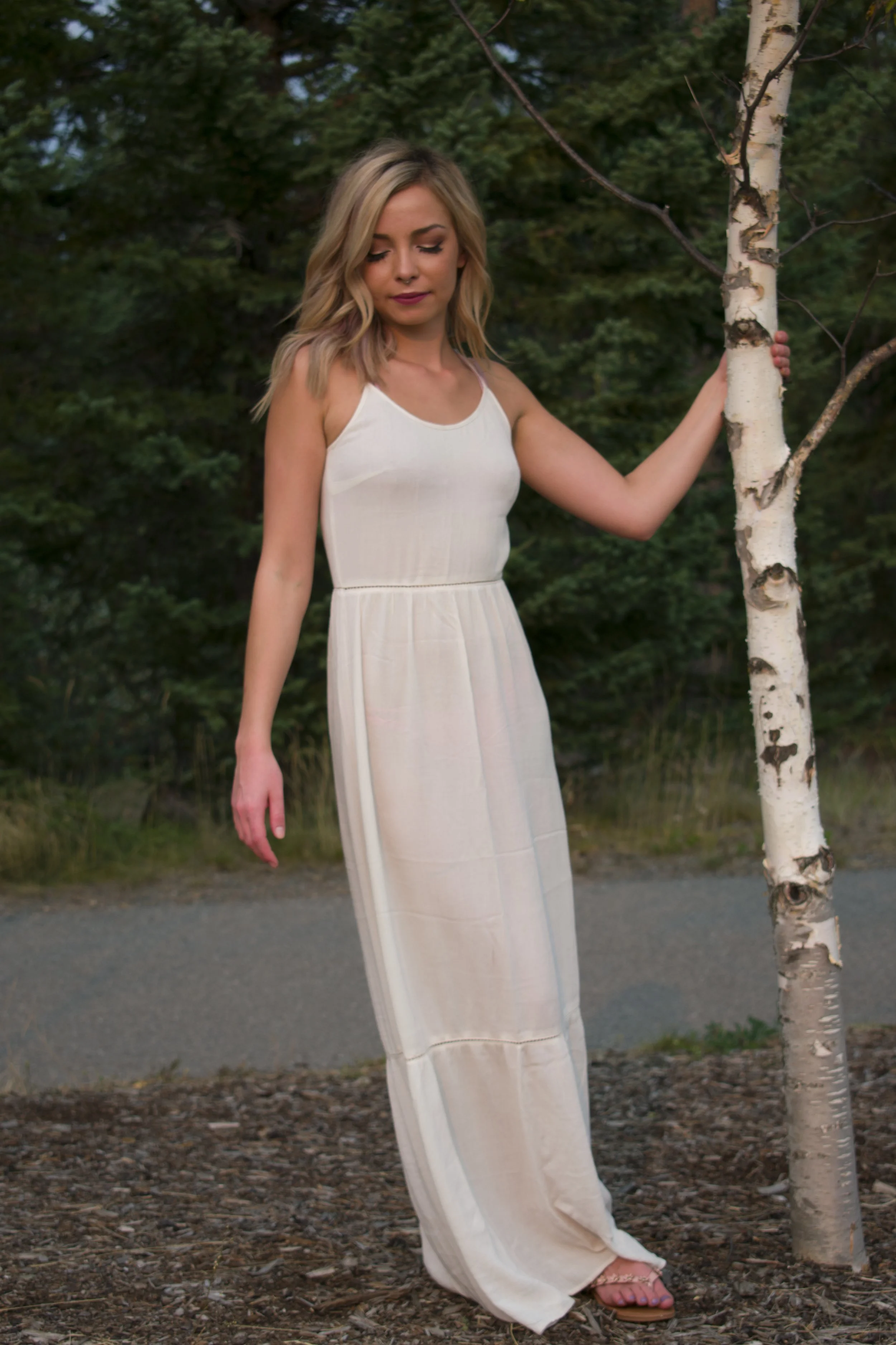 A young woman with blonde hair in a white sleeveless maxi dress standing outdoors near a white birch tree, surrounded by green trees and a dirt path.