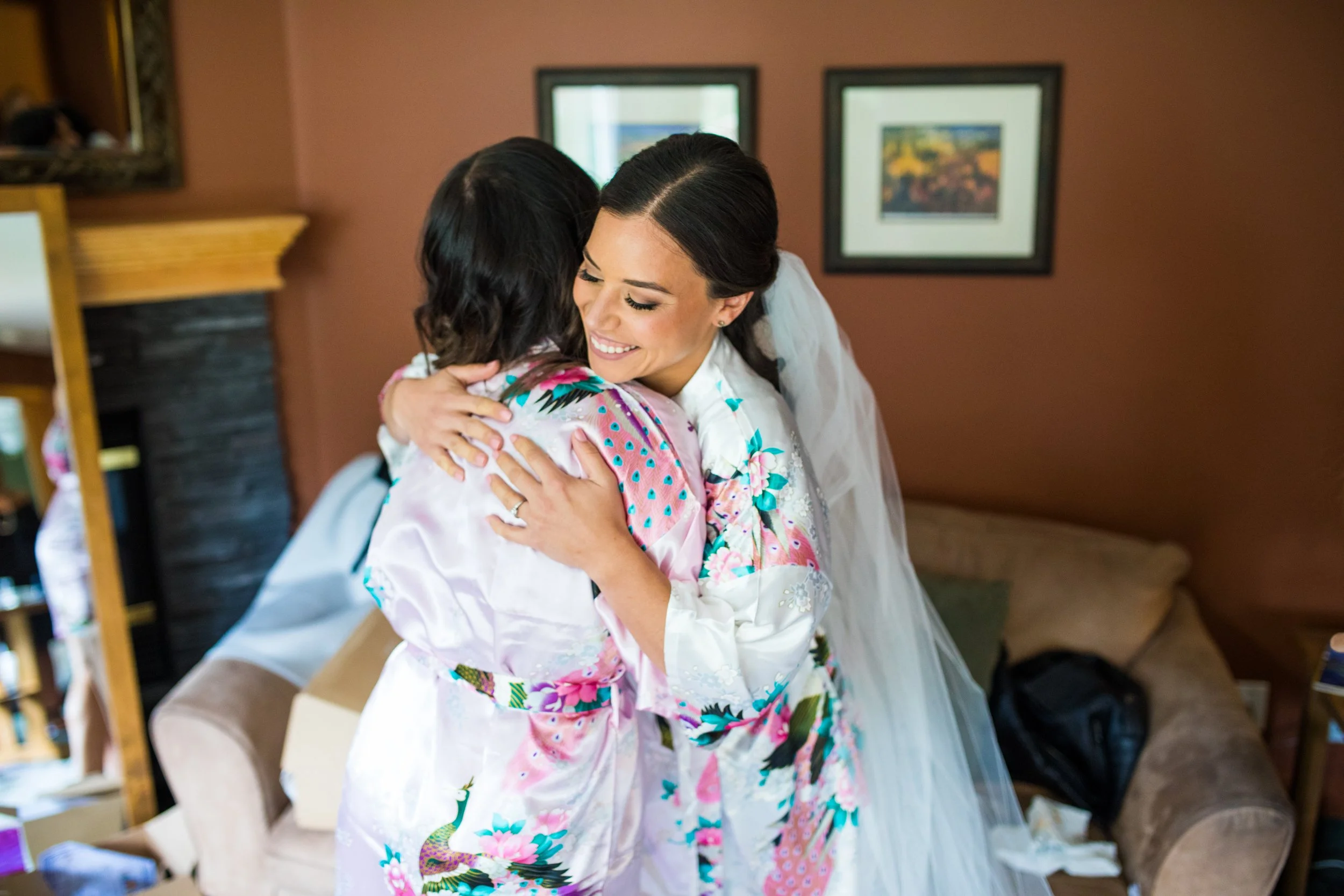 A bride in a floral and colorful wedding robe hugging another woman in a silk robe, inside a cozy room with a brown wall and framed artwork.