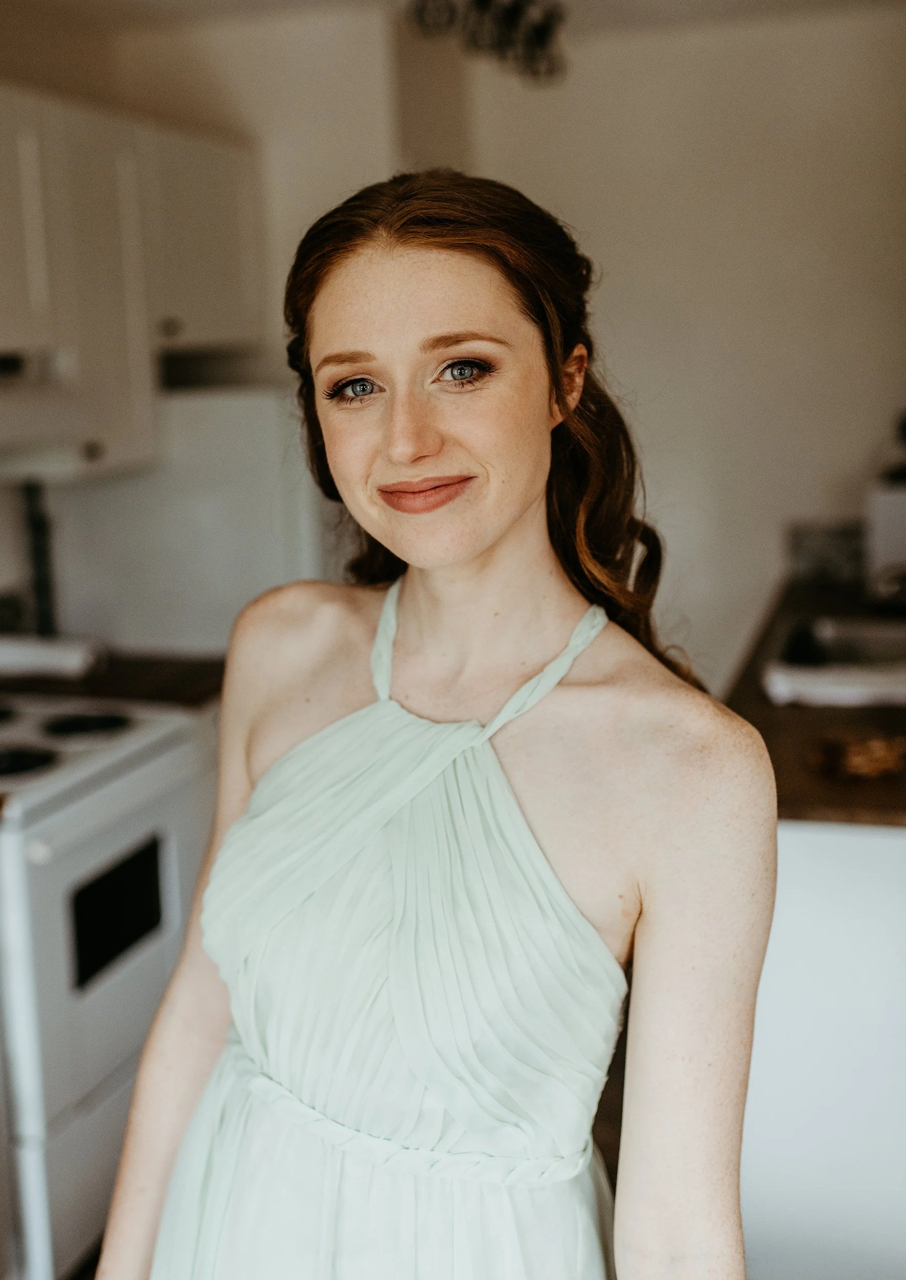 A woman with red hair, blue eyes, and freckles smiling in a kitchen, wearing a light green pleated dress.