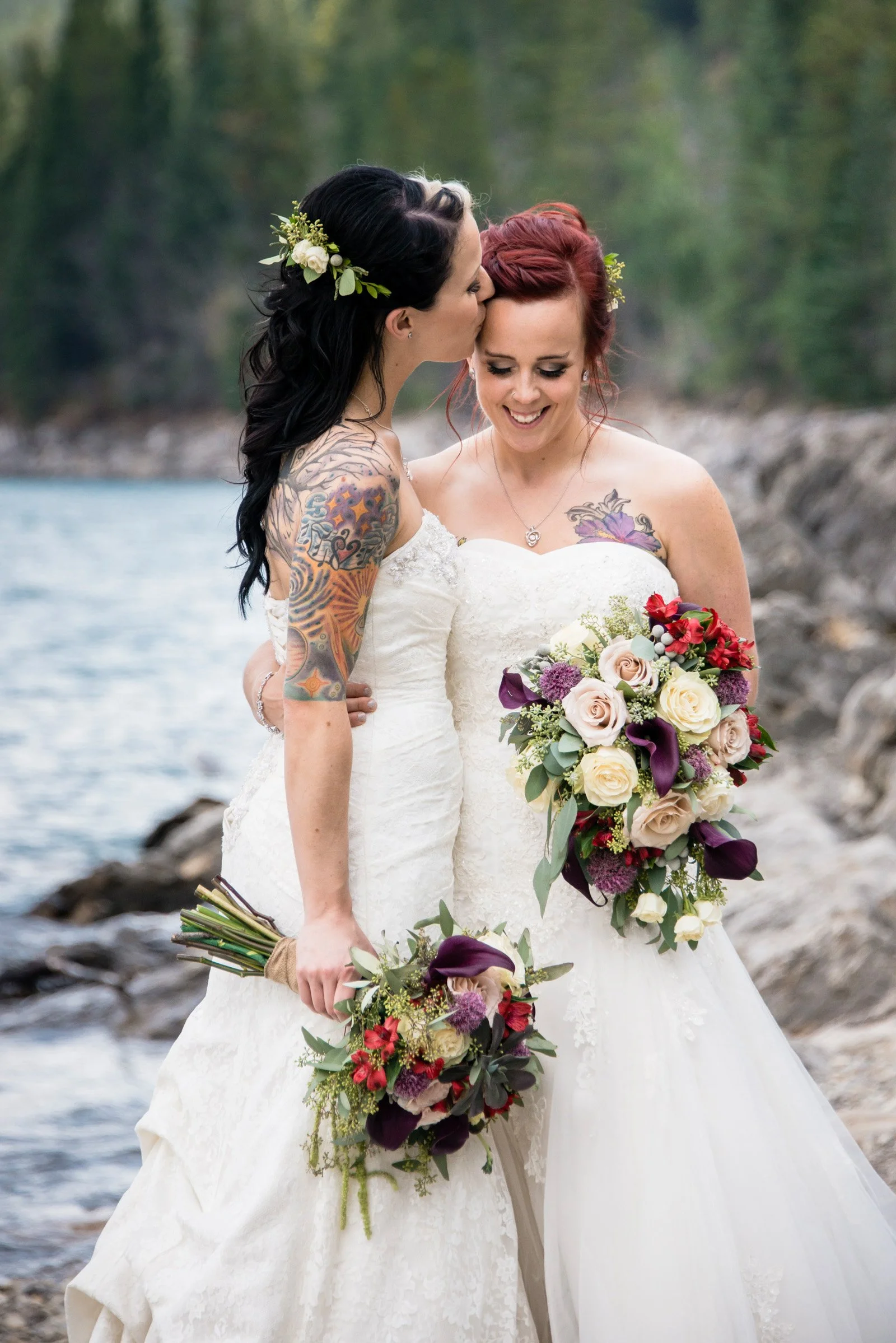 Two women in wedding dresses are standing close together outdoors near a body of water, holding bouquets of flowers, with one woman kissing the other's forehead.