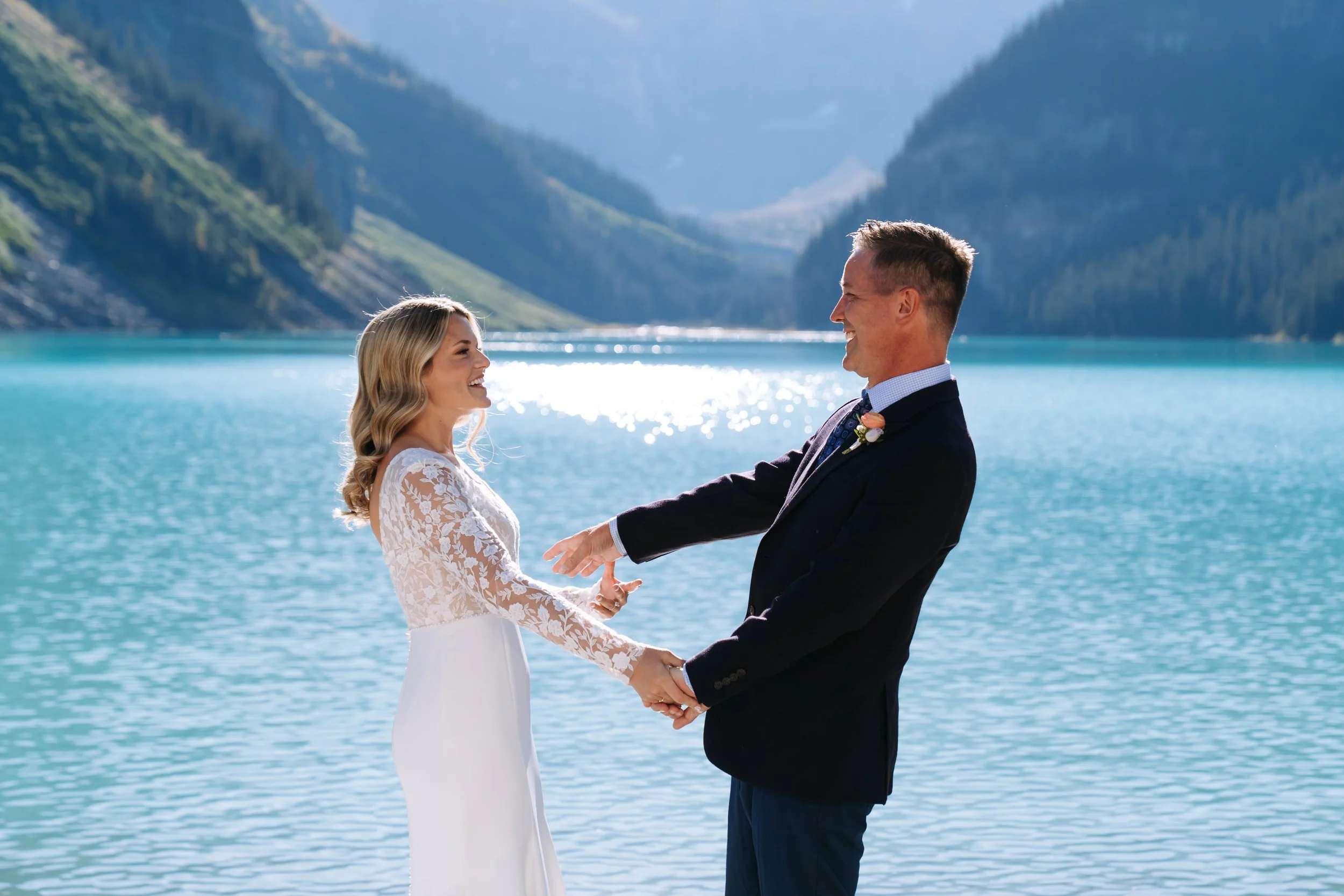 A bride and groom holding hands and smiling at each other by a lake with mountains in the background during their wedding.