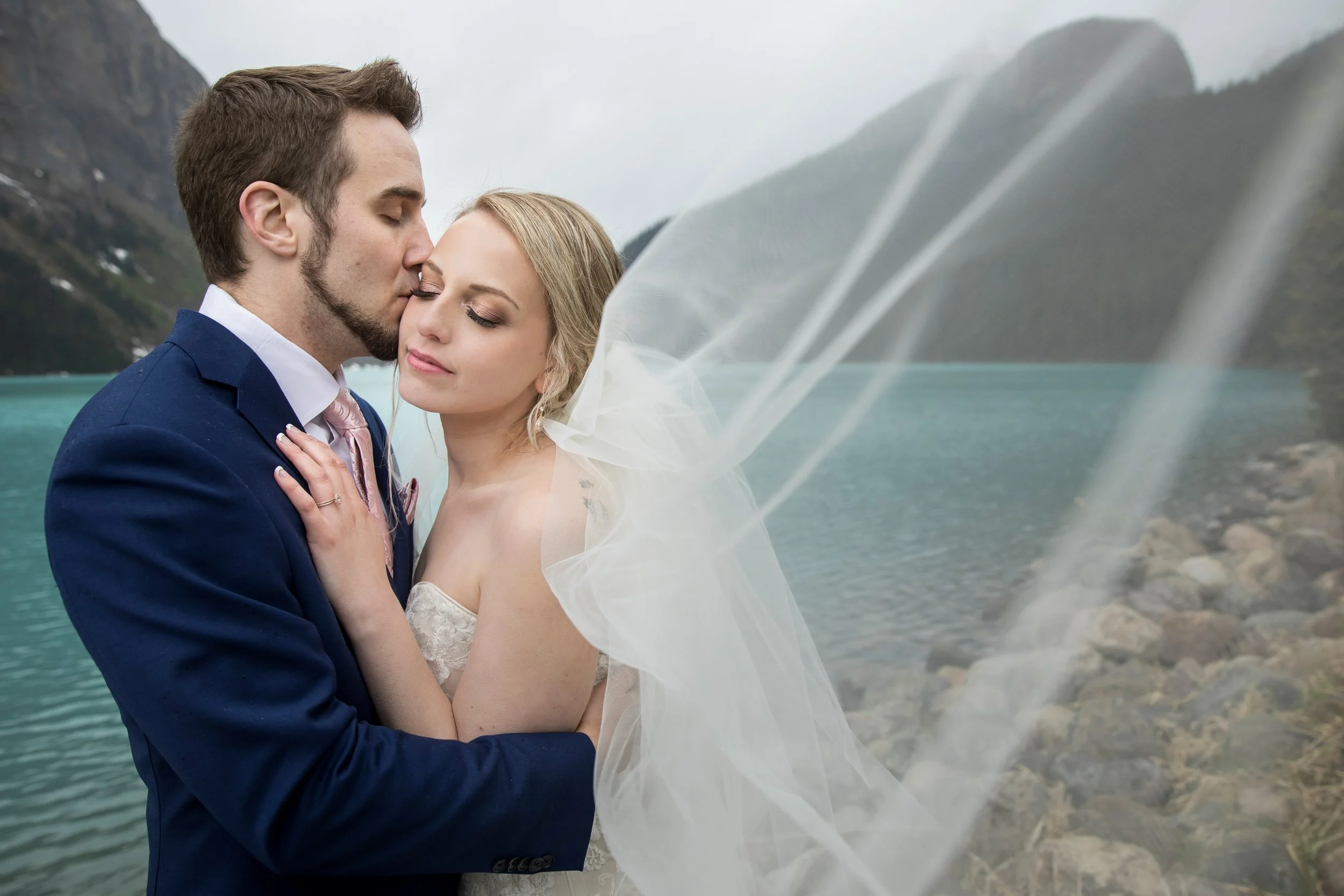 A bride and groom embracing near a lake with mountains in the background, the groom kissing the bride on the temple, and her eyes closed peacefully.