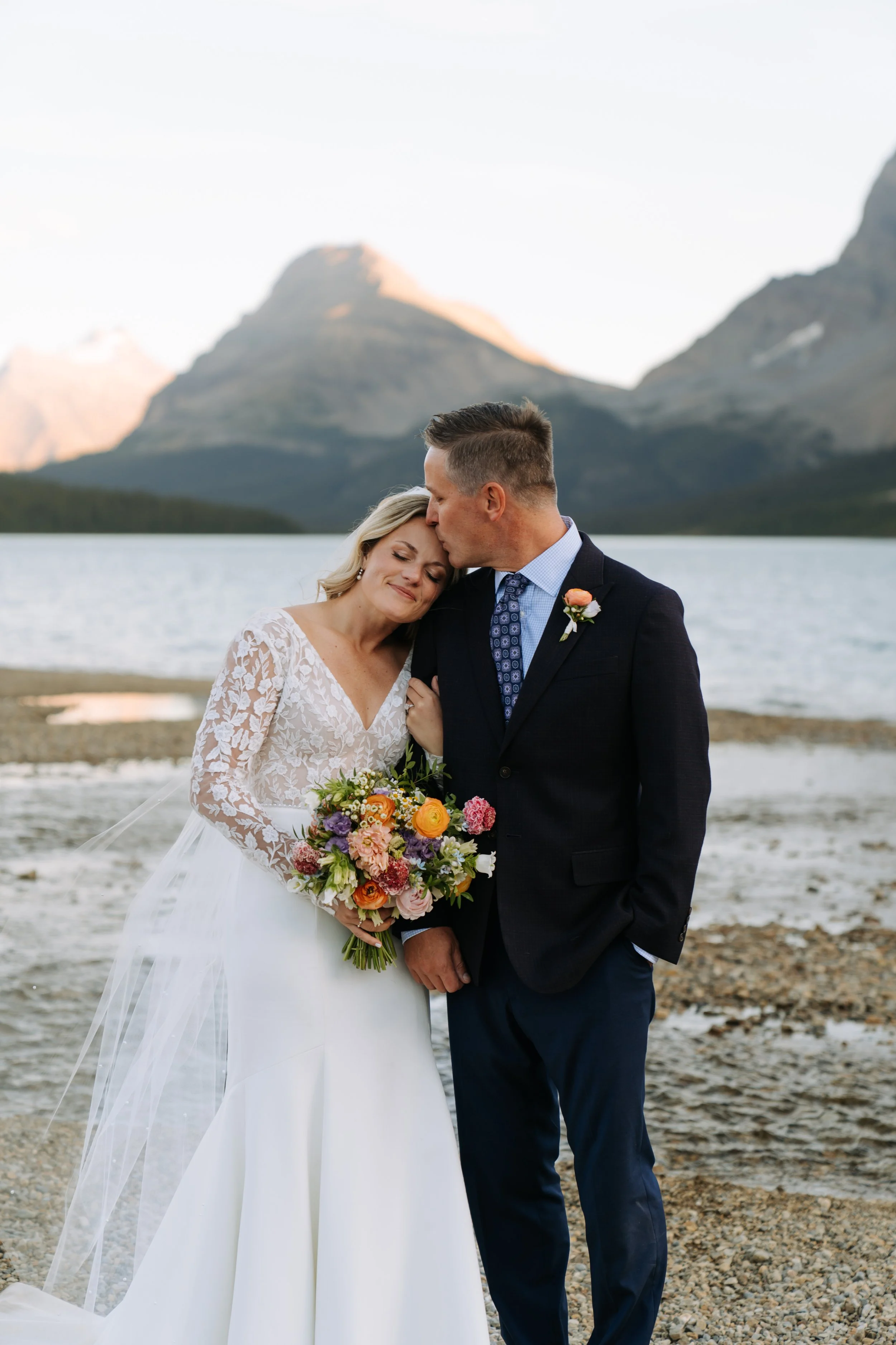 A newlywed couple standing on a lakeshore, with mountains in the background. The bride is holding a vibrant bouquet and leaning into the groom, who kisses her on the forehead.