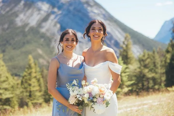 Two women in dresses holding bouquets, standing outdoors with mountains and trees in the background.