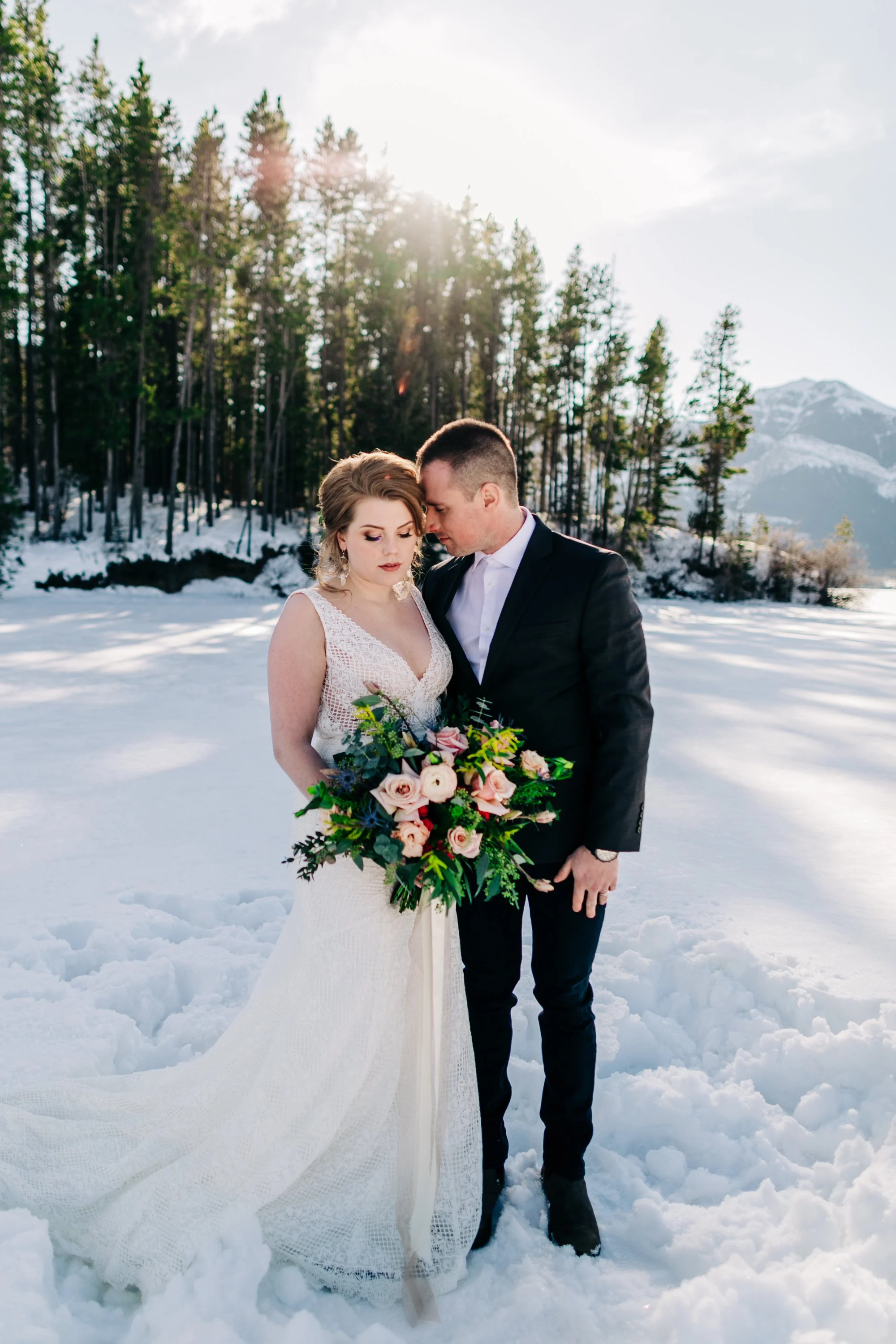 A bride and groom standing closely together on snow-covered ground in a winter landscape with trees and mountains in the background. The bride is holding a large bouquet of flowers and is wearing a white wedding dress, while the groom is dressed in a