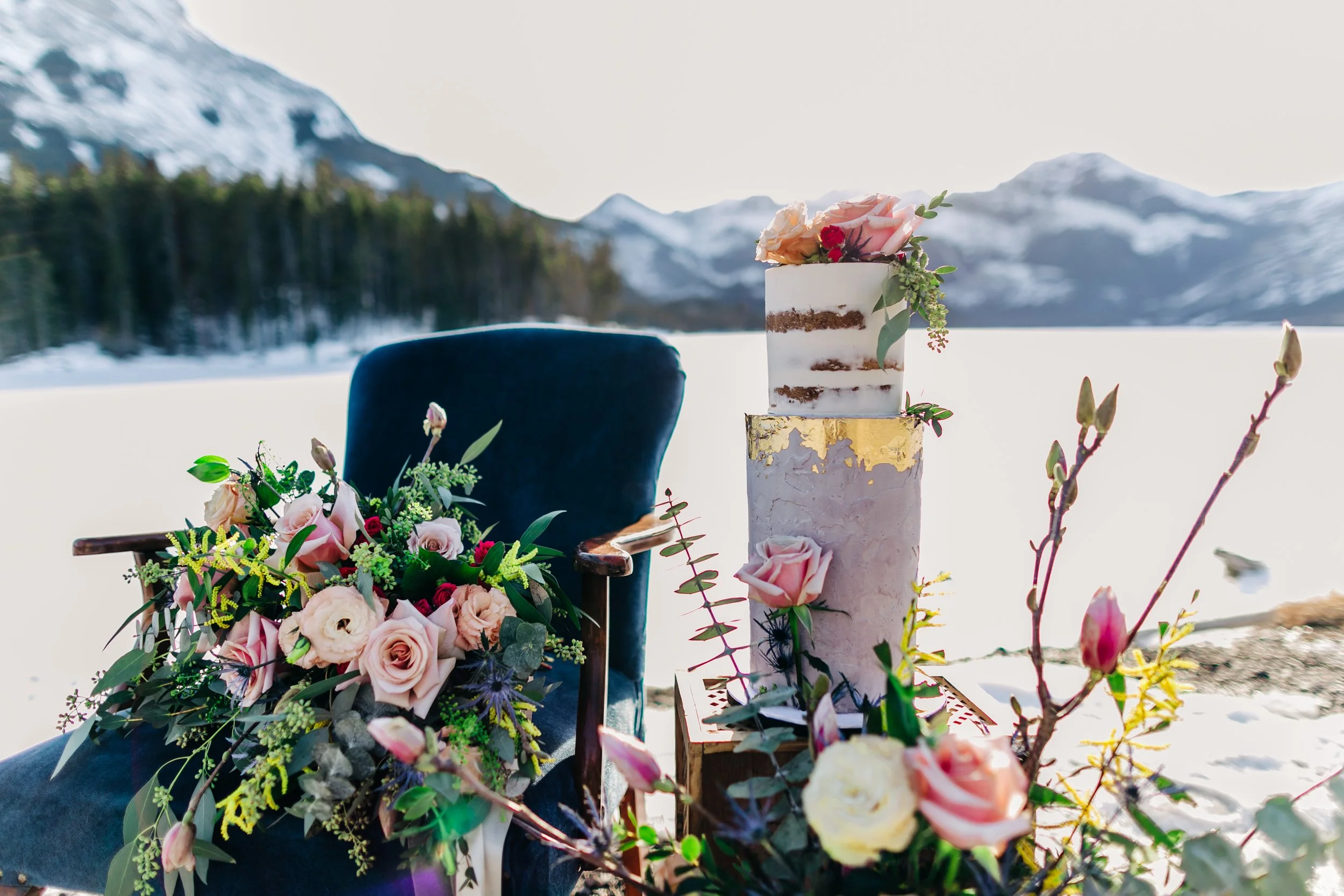 A wedding setup outdoors in a snowy landscape with mountains in the background. There is a dark blue velvet chair adorned with a floral arrangement of pink, white, and red roses and greenery. Next to the chair is a tall, decorated cake with a gold le