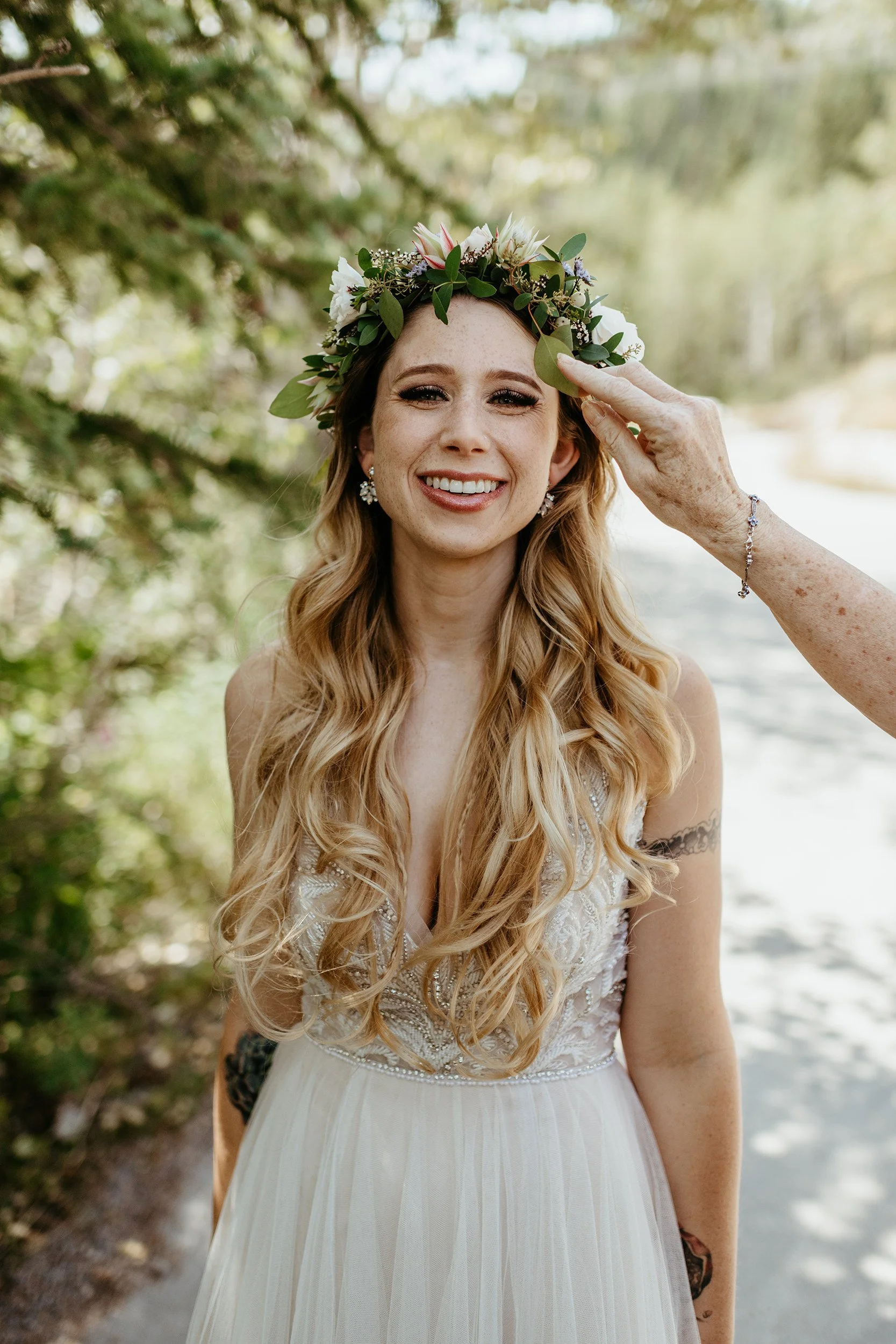 Bride with long, wavy blond hair wearing a white wedding dress and a floral crown, smiling outdoors with greenery and water in the background.
