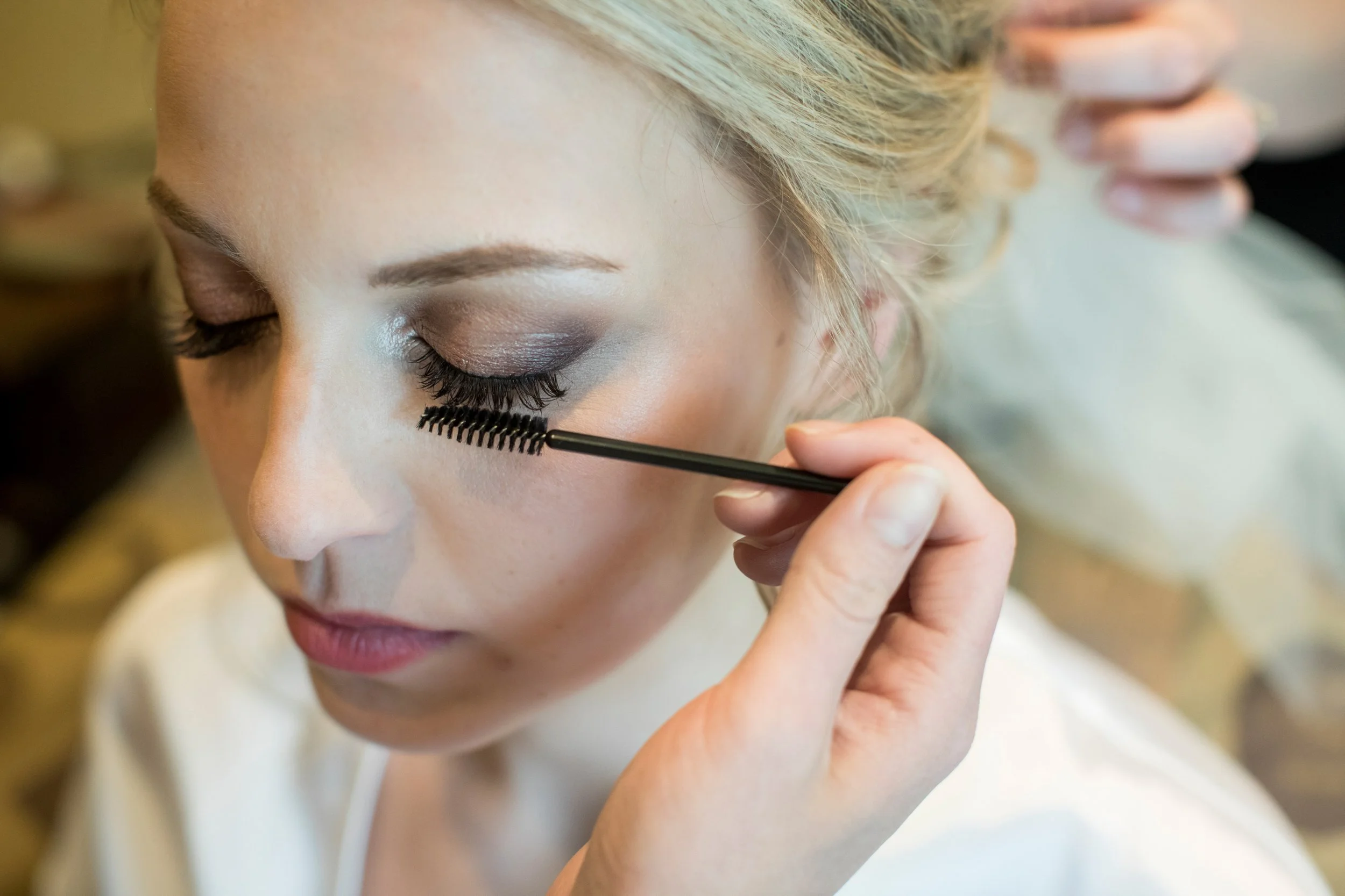 A woman with makeup being applied around her eye area, with her eyes closed, by someone using a mascara brush.