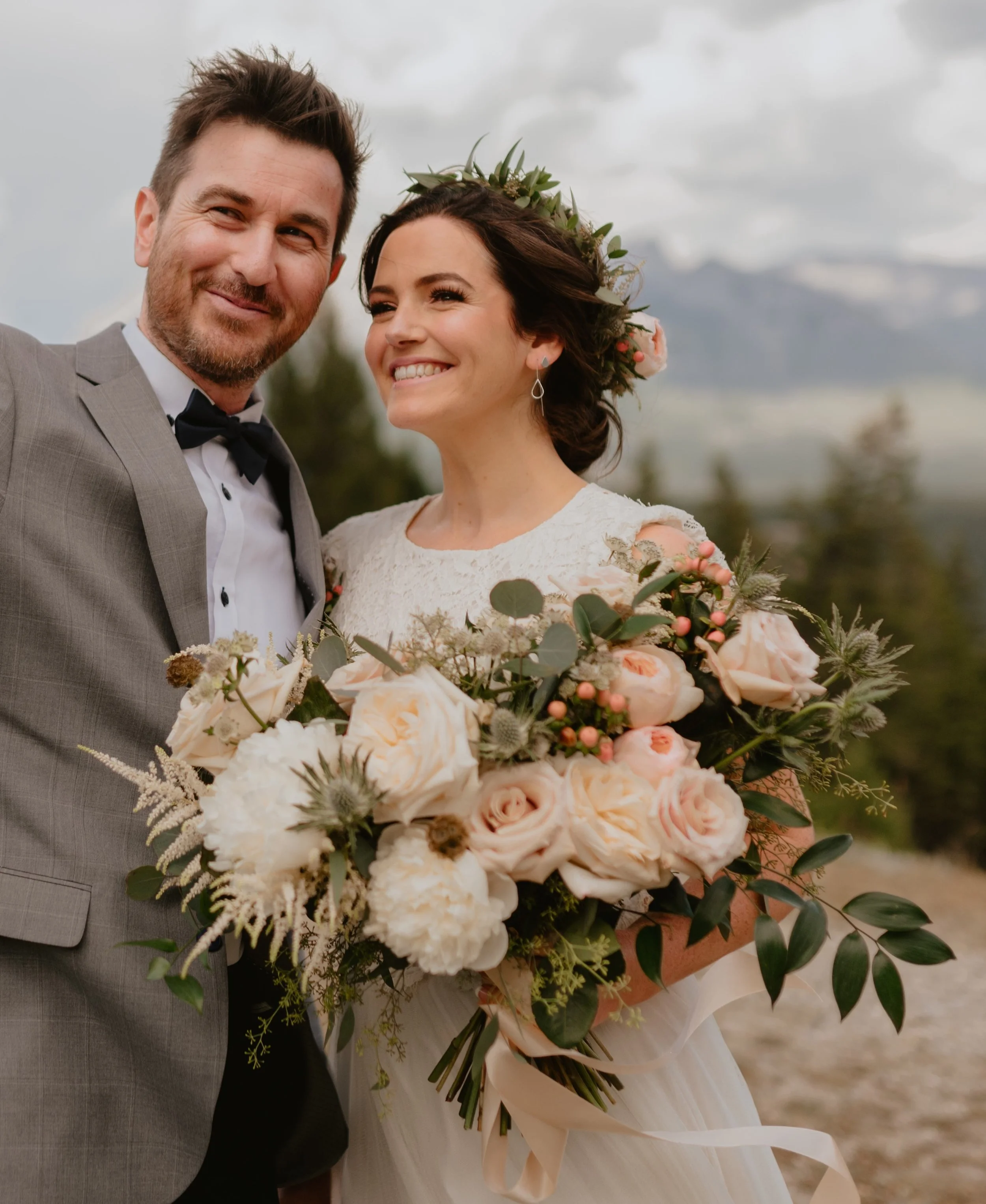 A newlywed couple taking a selfie outdoors, with the bride holding a large bouquet of pink and white roses, greenery, and other flowers. The woman wears a white wedding dress and a floral crown, and the man wears a gray suit with a black bow tie. The